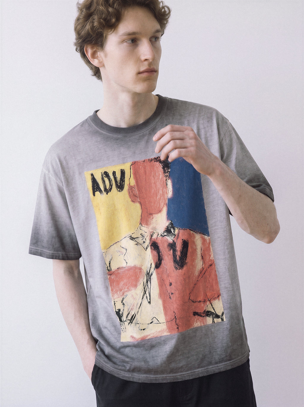 A male model touching the hem wearing an ombre grey faceless portrait oil pastel graphic tee, capturing the visual texture and casual drape.