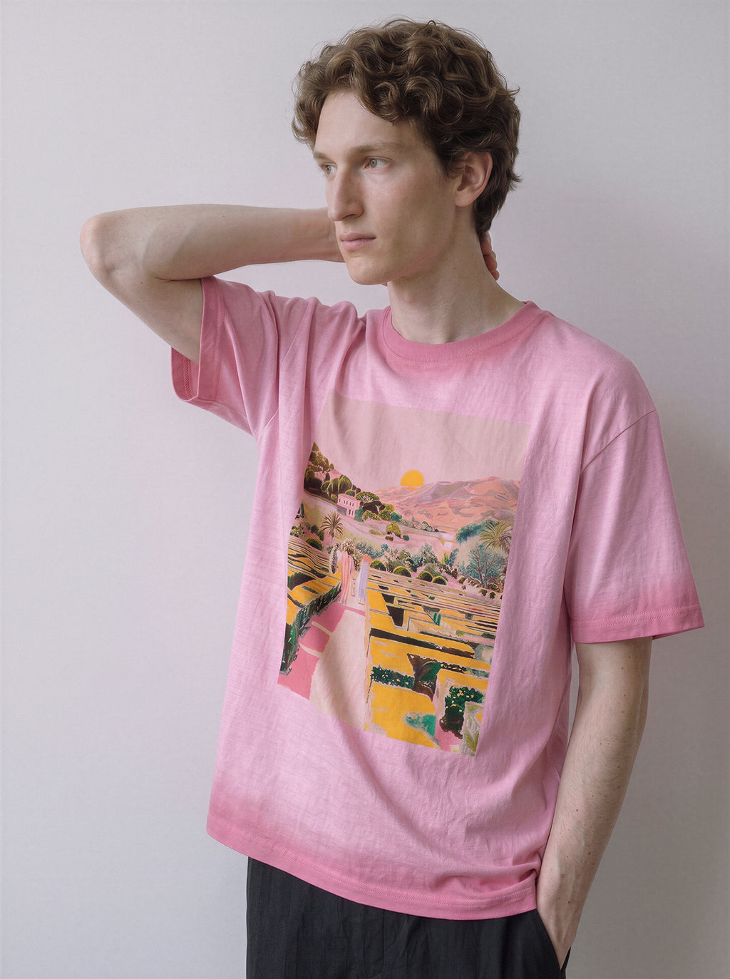A male model with his hand behind his neck wearing an aura pink geometric labyrinth graphic novel tee, displaying the geometric maze design and casual drape.