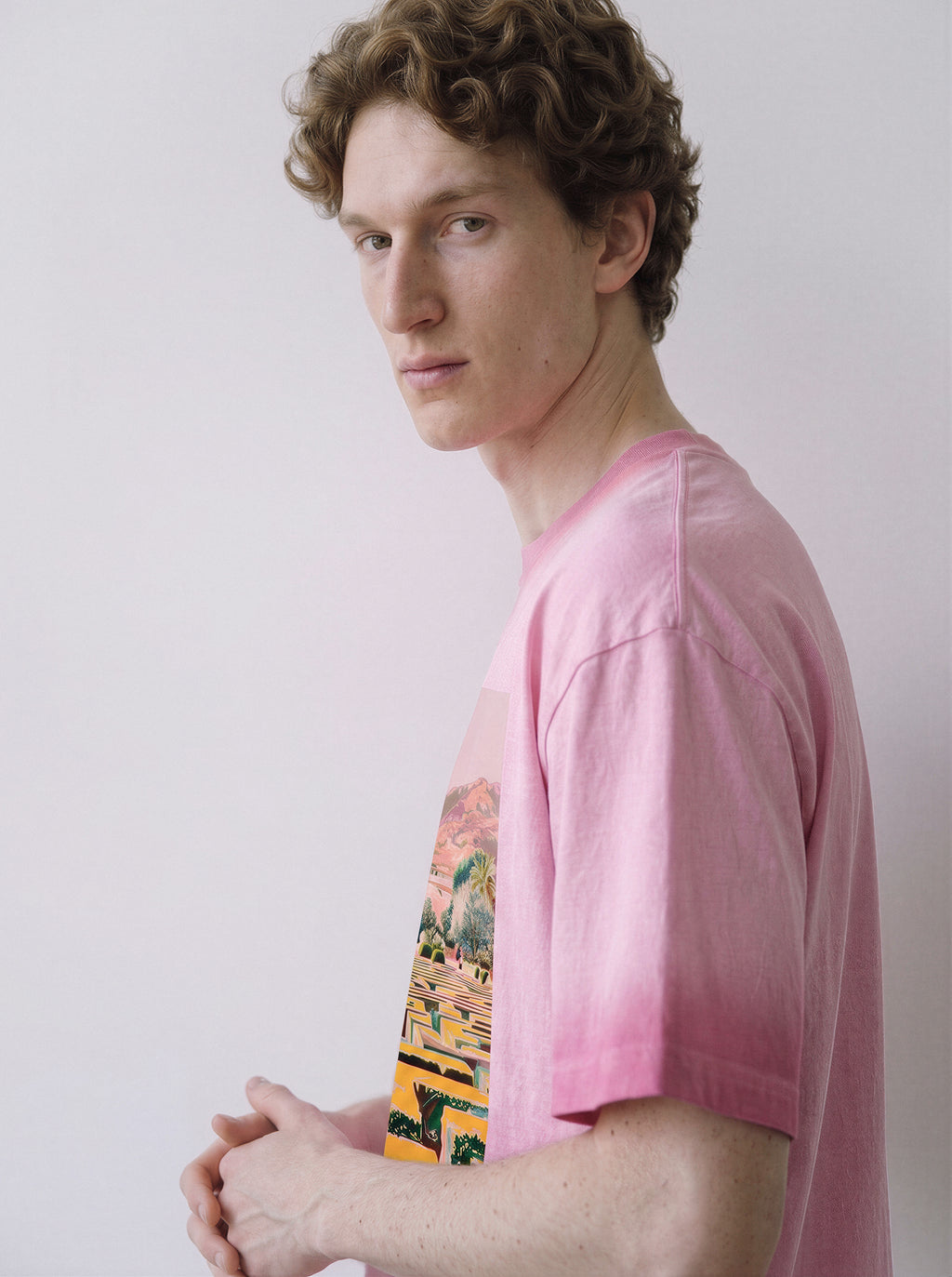 A side profile of a male model looking at the camera wearing an aura pink geometric labyrinth graphic novel tee, emphasizing the surreal landscape graphic and unwritten graphic novel aesthetic.