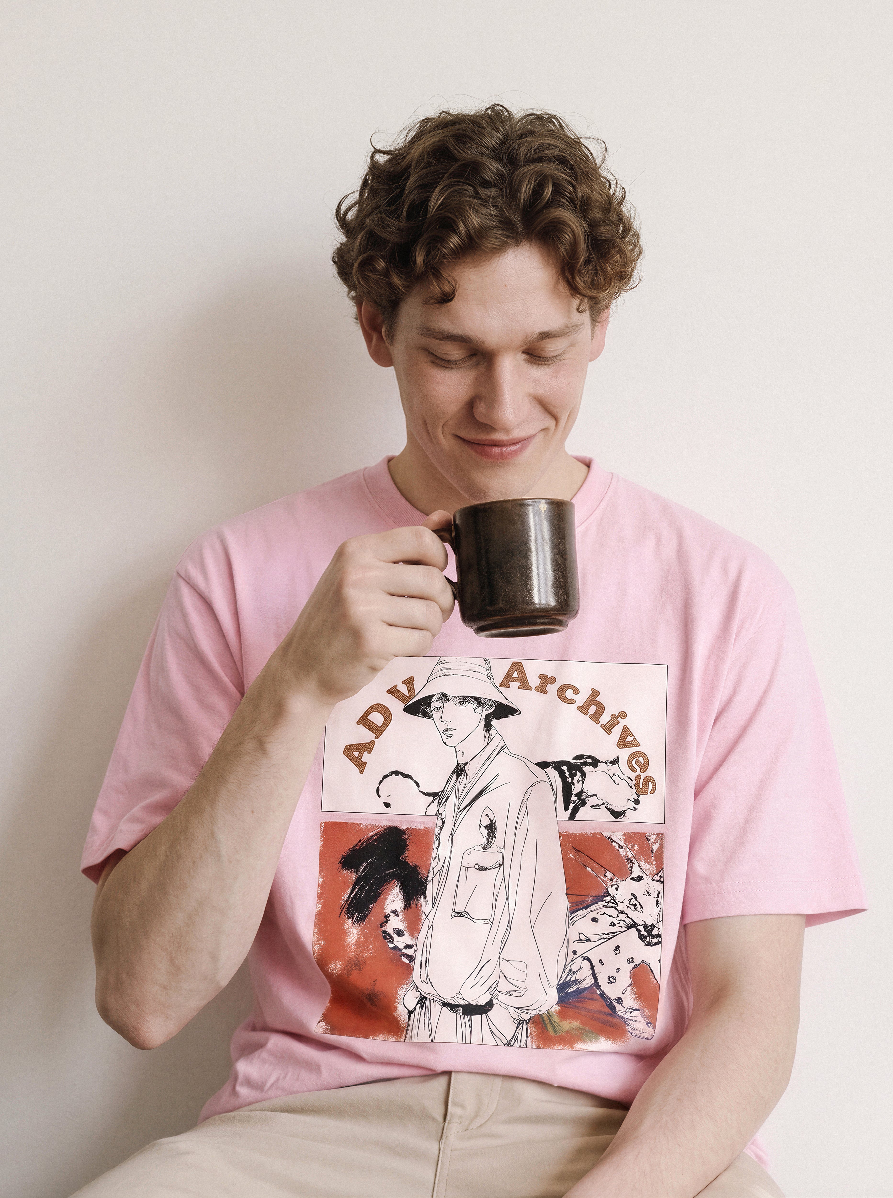A seated male model bringing a mug to his mouth wearing an aura pink graphic novel boy and wildlife tee, emphasizing the paper wilderness comic illustration style.