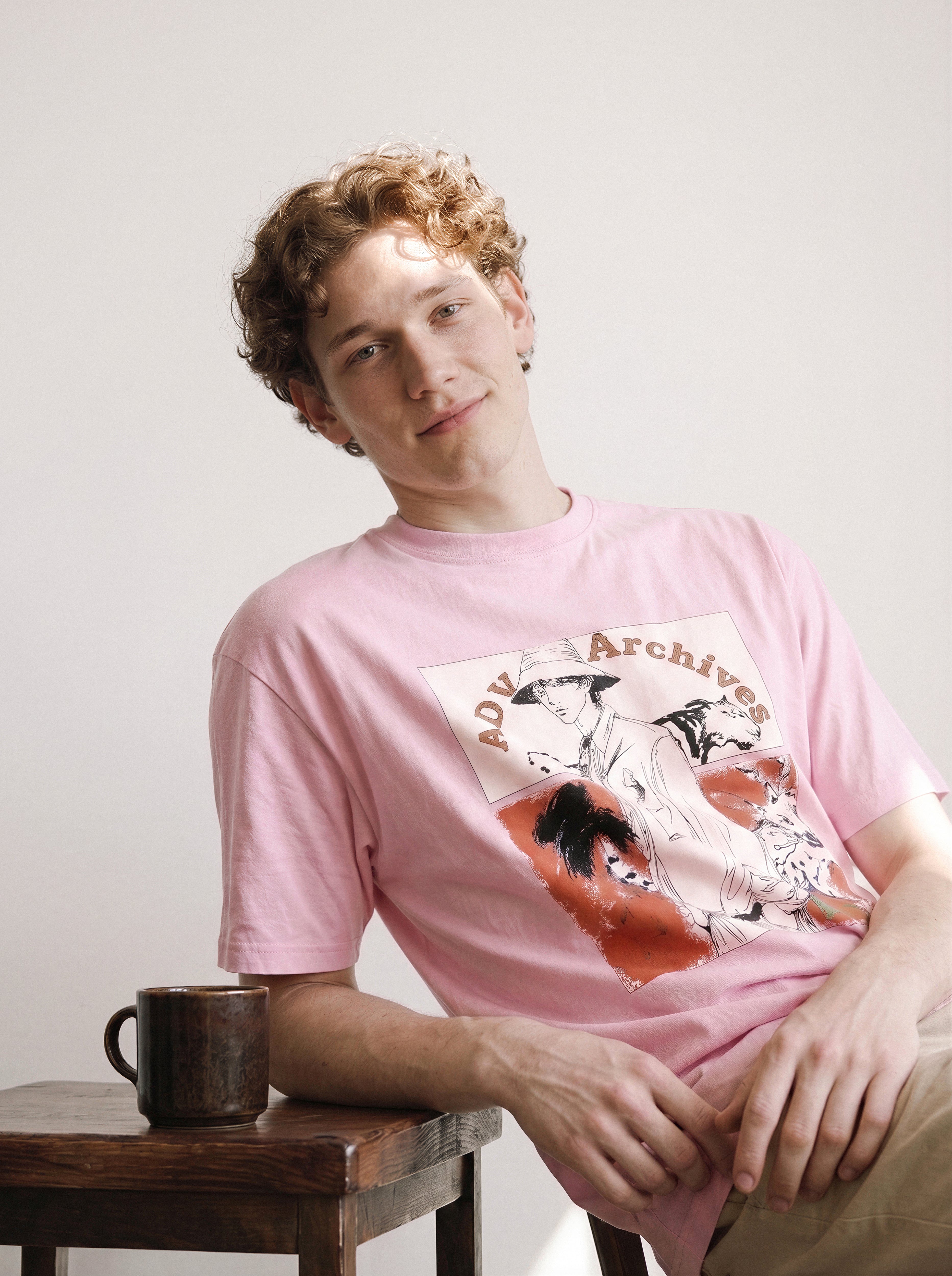 A male model leaning on a wooden stool next to a mug wearing an aura pink graphic novel boy and wildlife tee, displaying the casual retro graphic novel art and vintage streetwear vibe.