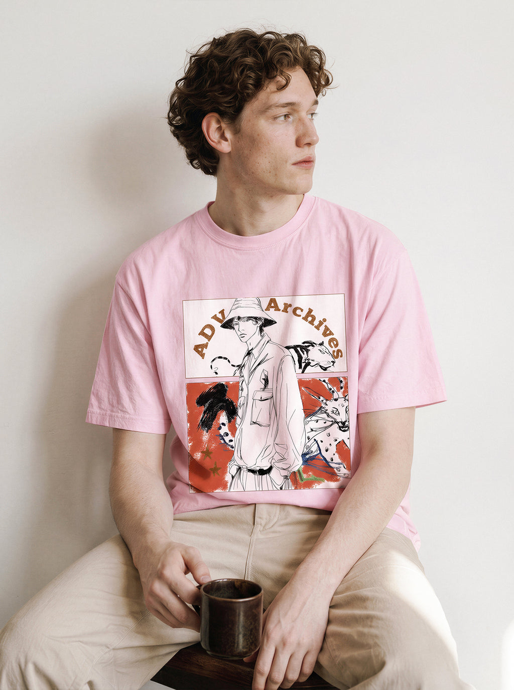 A seated male model looking to the side holding a mug wearing an aura pink graphic novel boy and wildlife tee, capturing the unfinished comic panel graphic design.