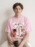 A seated male model holding a mug and looking at the camera wearing an aura pink graphic novel boy and wildlife tee, showcasing the vintage comic panel print and relaxed boxy fit.