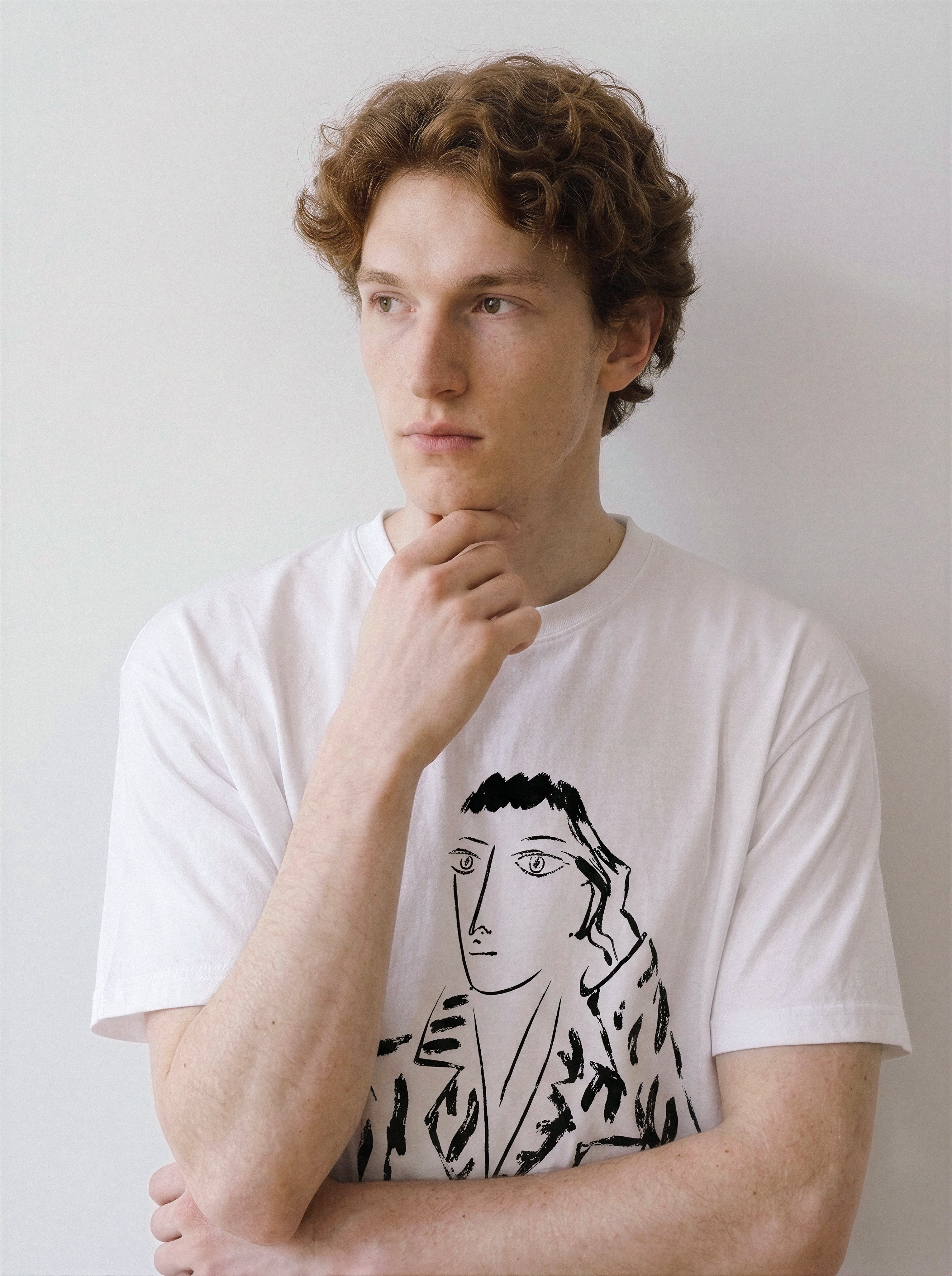 A male model resting his hand on his chin wearing a white hand-lettered dream portrait line art graphic tee, showcasing the high-contrast pure charcoal sketch illustration.