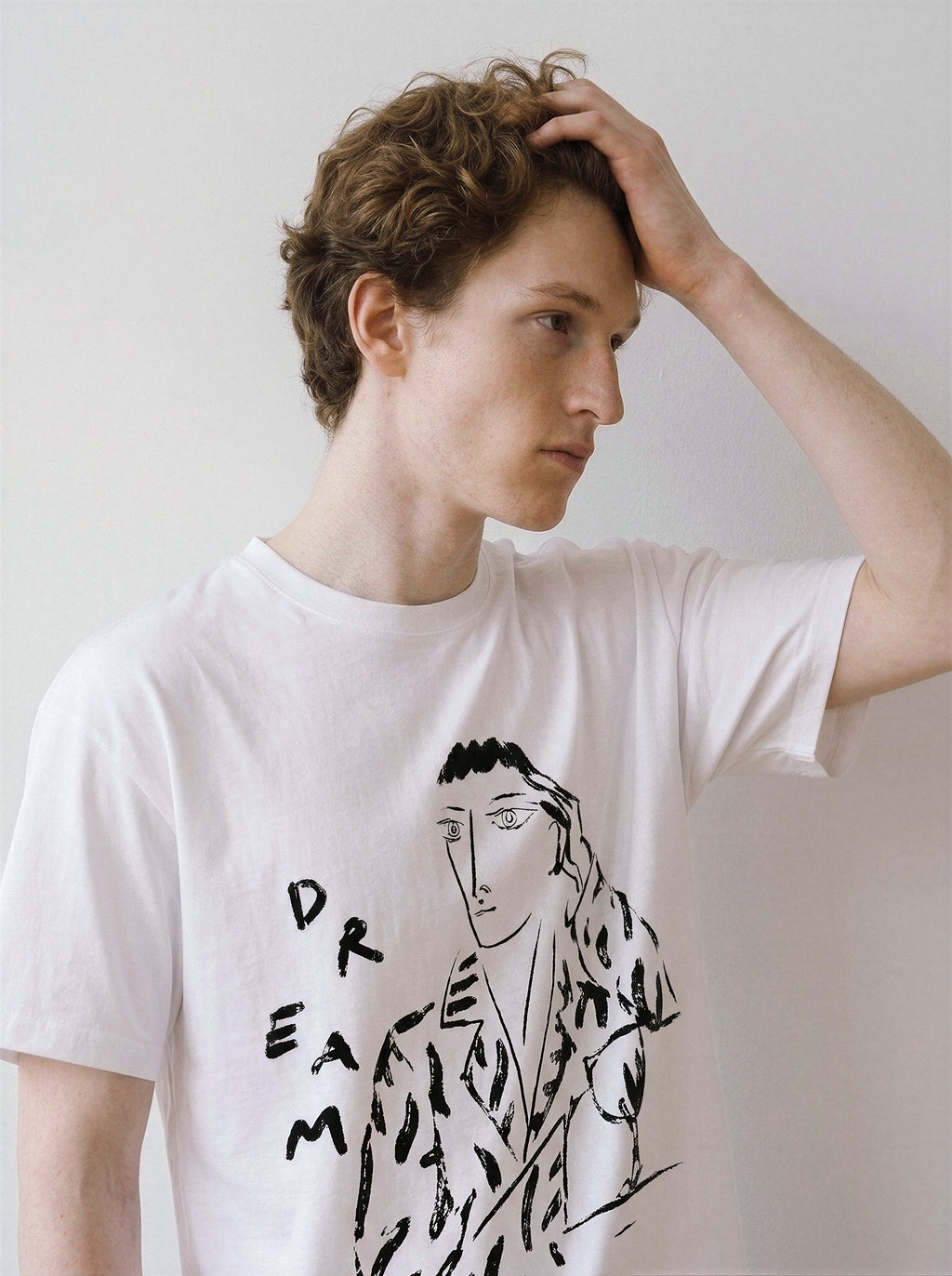 A male model with his hand in his hair looking away, wearing a white hand-lettered dream portrait line art graphic tee, demonstrating the edgy line art streetwear style.
