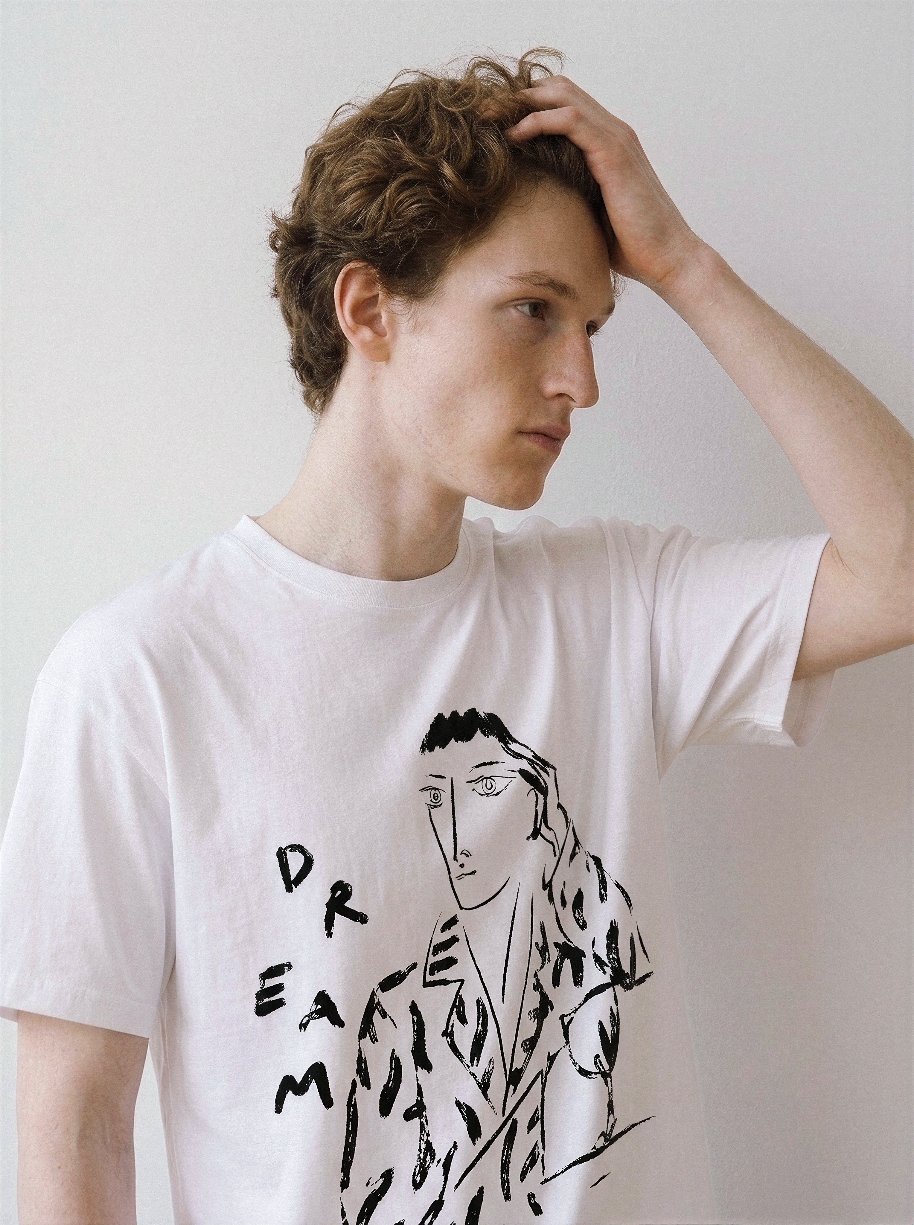 A male model with his hand in his hair looking away, wearing a white hand-lettered dream portrait line art graphic tee, demonstrating the edgy line art streetwear style.