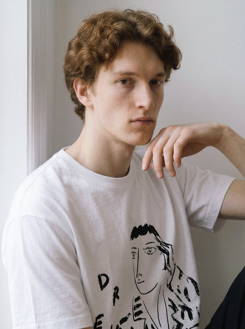 A male model sitting and resting his arm while wearing a white hand-lettered dream portrait line art graphic tee, emphasizing the relaxed unisex fit and raw ink texture.