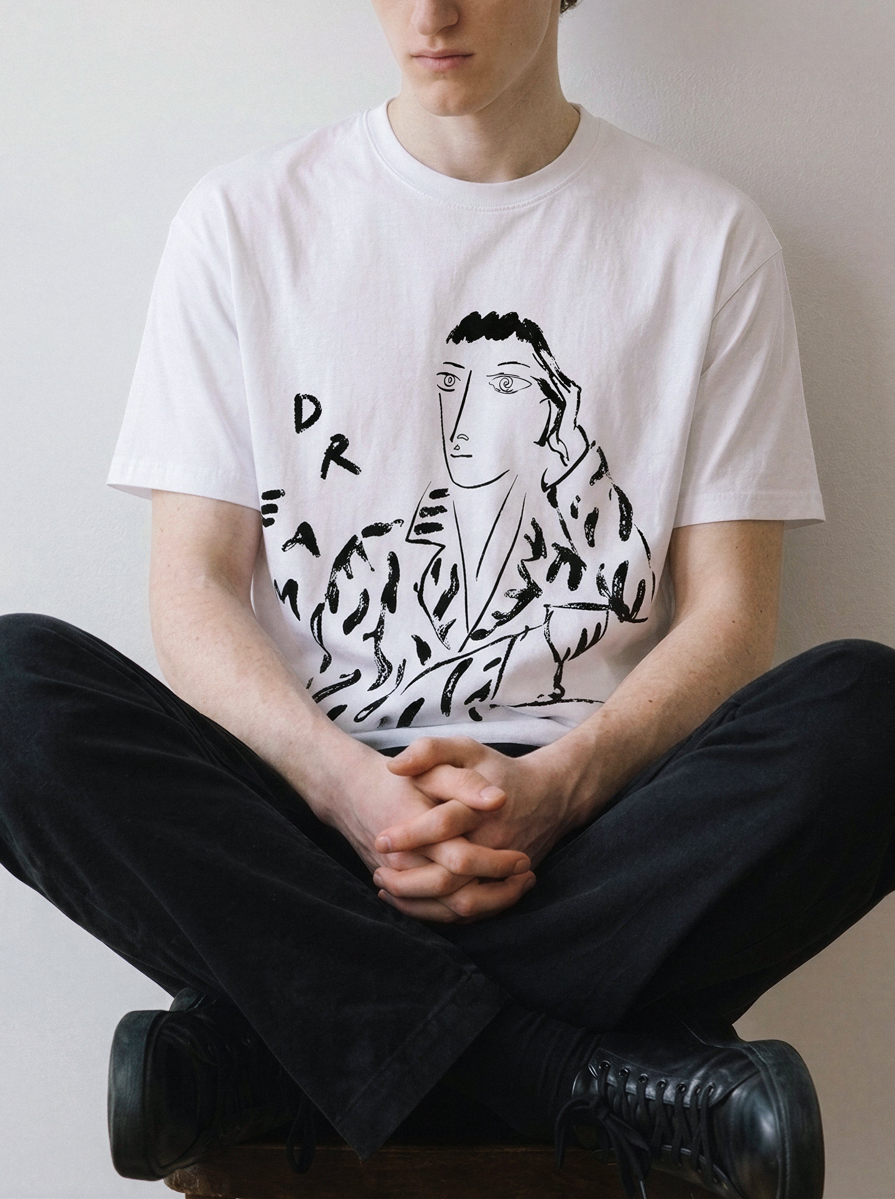 A male model sitting cross-legged wearing a white hand-lettered dream portrait line art graphic tee, capturing the minimalist streetwear look and striking typography graphic.