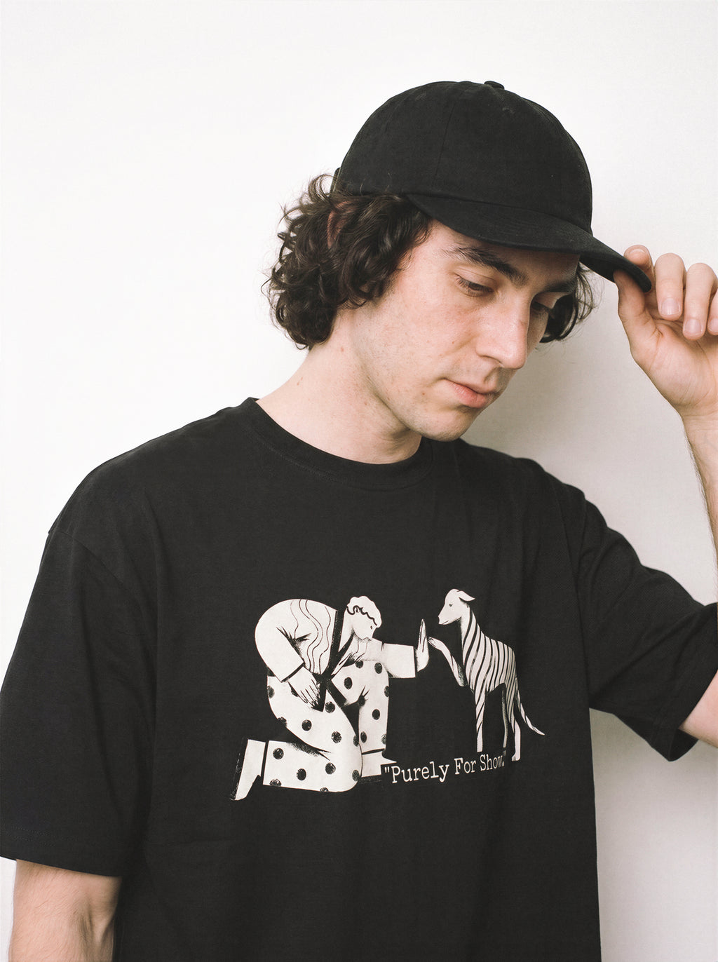 A male model holding his cap and looking right wearing a black handshake gesture line art graphic tee, emphasizing the exhausted by fakeness aesthetic and casual drape.