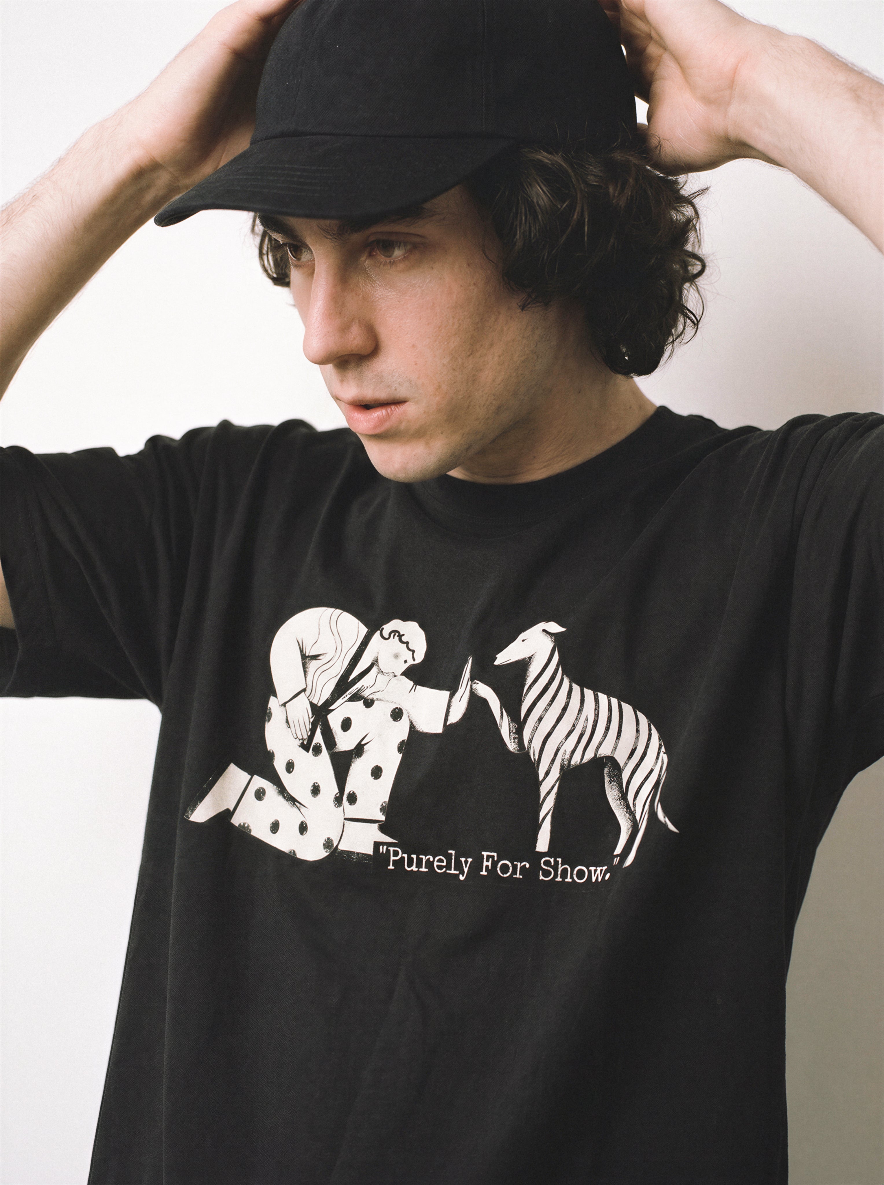 A male model touching his cap and looking left wearing a black handshake gesture line art graphic tee, highlighting the purely for show mindset and relaxed boxy fit.