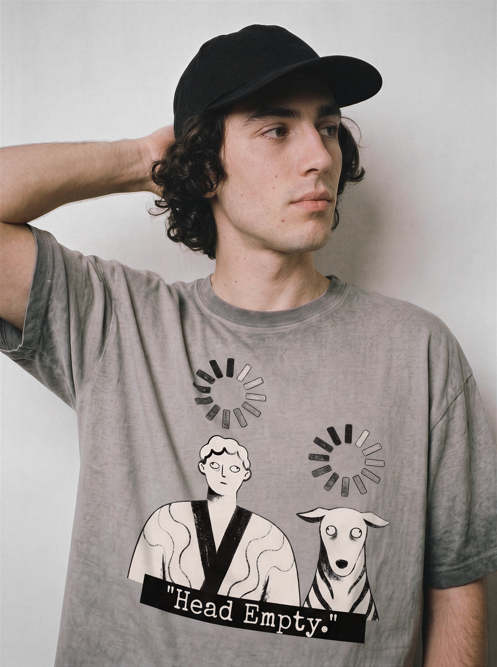 A male model with his hand behind his head wearing an ombre grey head empty buffering icon graphic tee, emphasizing the corporate burnout graphic and casual drape.