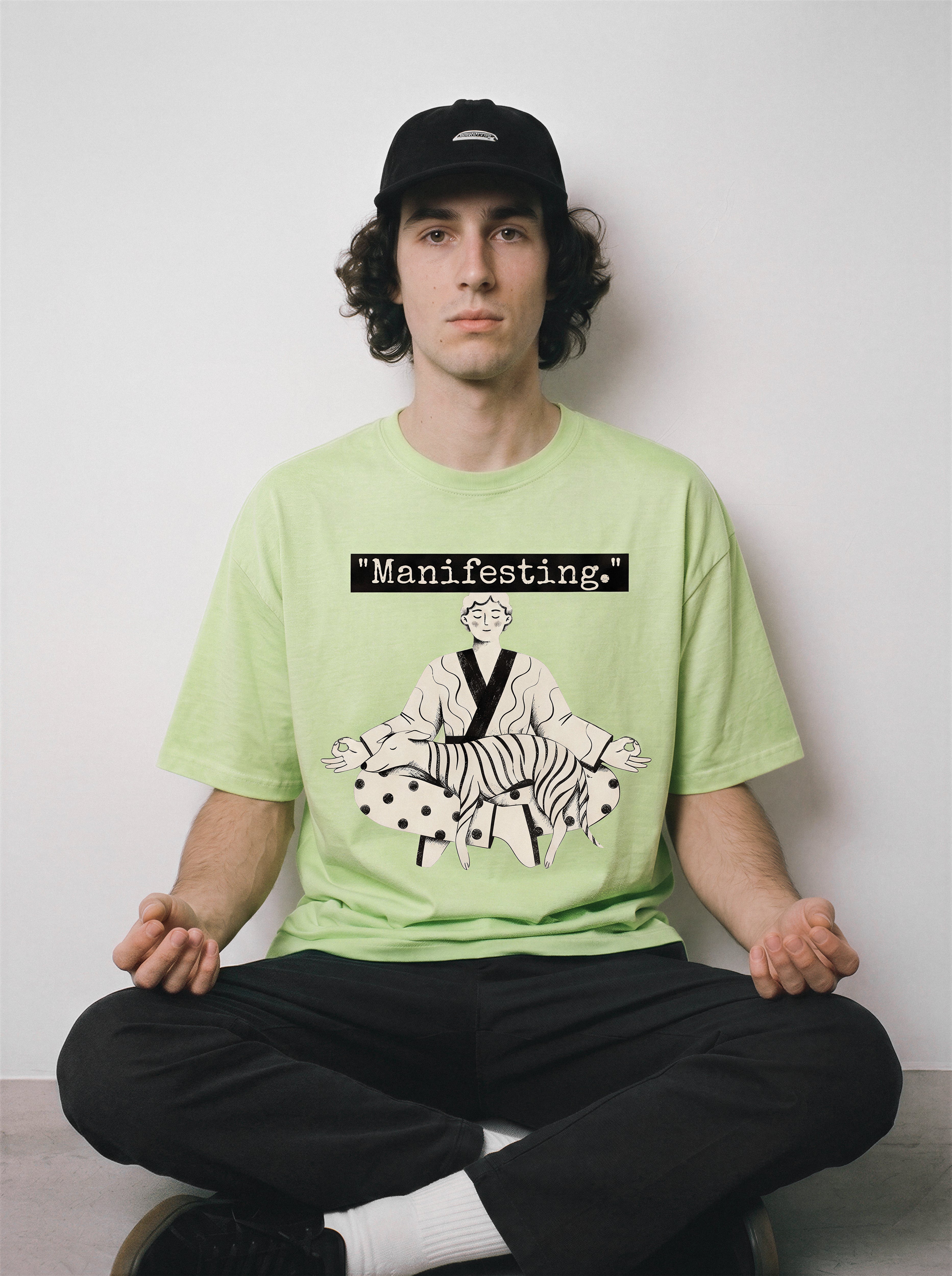 A male model sitting cross-legged wearing an acid lime manifesting meditating graphic tee, showcasing the existential deadpan boy and dog line art design.