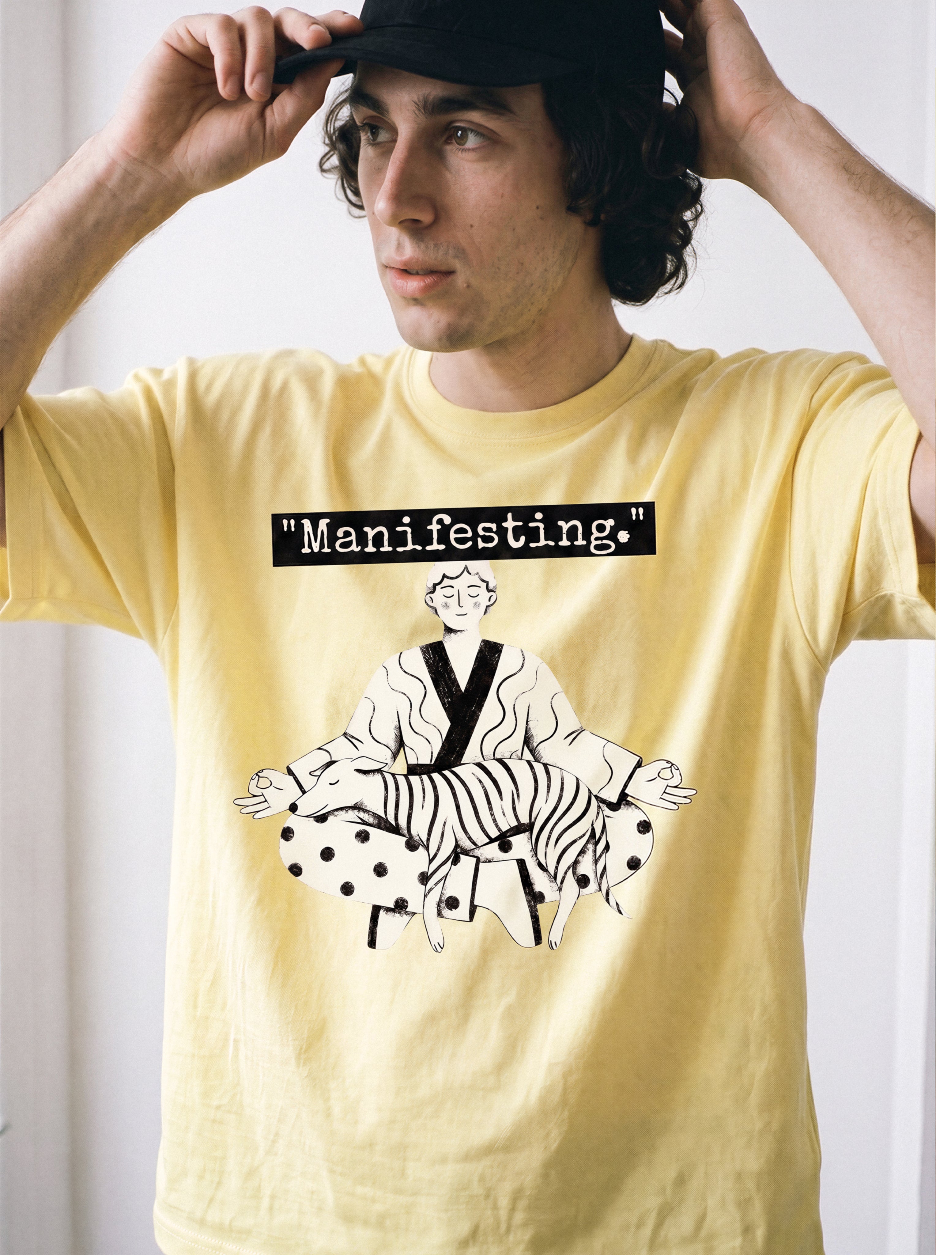 A male model adjusting his cap wearing a butter yellow meditating boy and dog line art graphic tee, showcasing the manifesting typography and independent manga aesthetic.