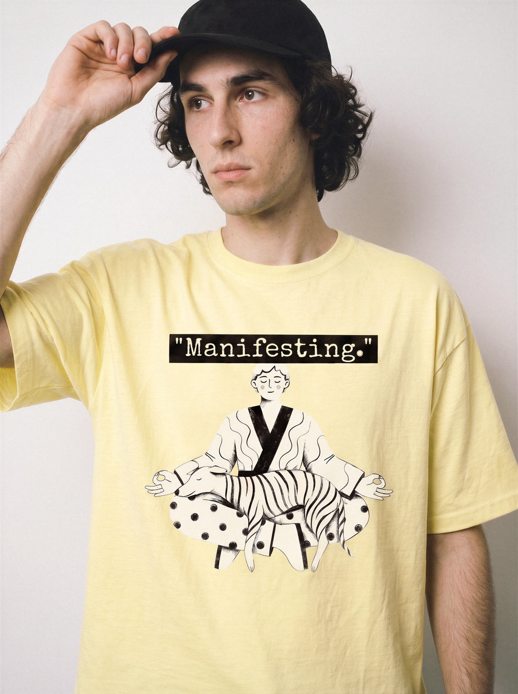 A front view of a male model holding his cap wearing a butter yellow meditating boy and dog line art graphic tee, emphasizing the clean monochrome illustration and relaxed fit.