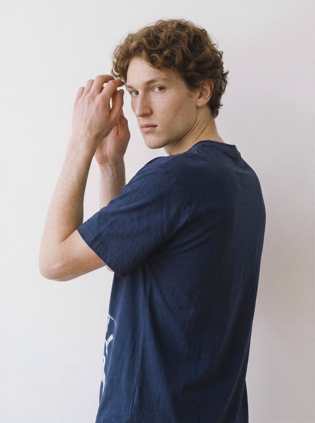 Side angle view of a male model with hands on his head, wearing a navy 180g oversized cotton tee with a striking unshaded chalk portrait print.