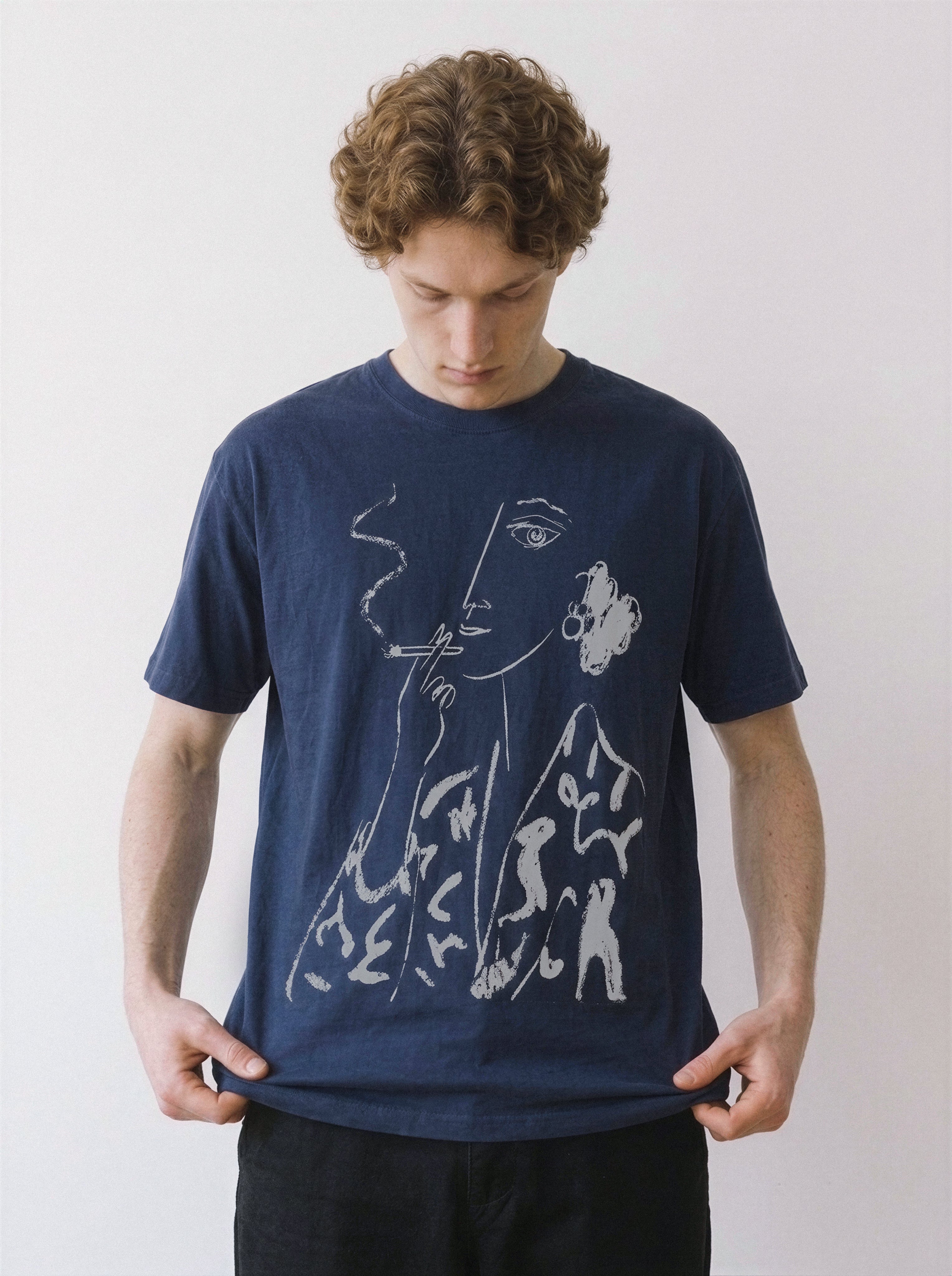 A man wearing a navy blue t-shirt with a white minimalist line art graphic of a face smoking, posed against a plain off-white background.