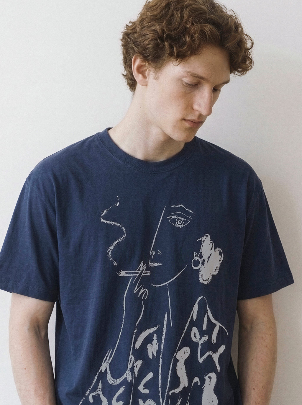 Front portrait of a male model looking down, wearing a navy oversized graphic tee featuring a chalk-textured smoking figure line art portrait.