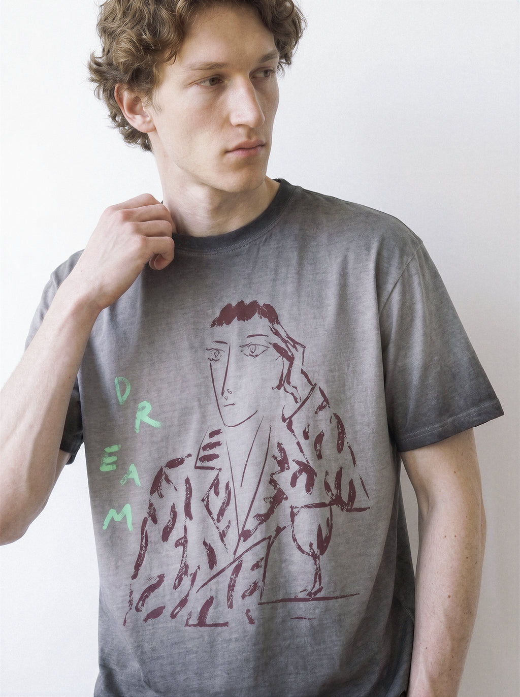 Front portrait of a male model looking away and adjusting his collar, wearing an ombre grey heavyweight boxy fit graphic tee featuring a raw unshaded charcoal portrait and DREAM typography.