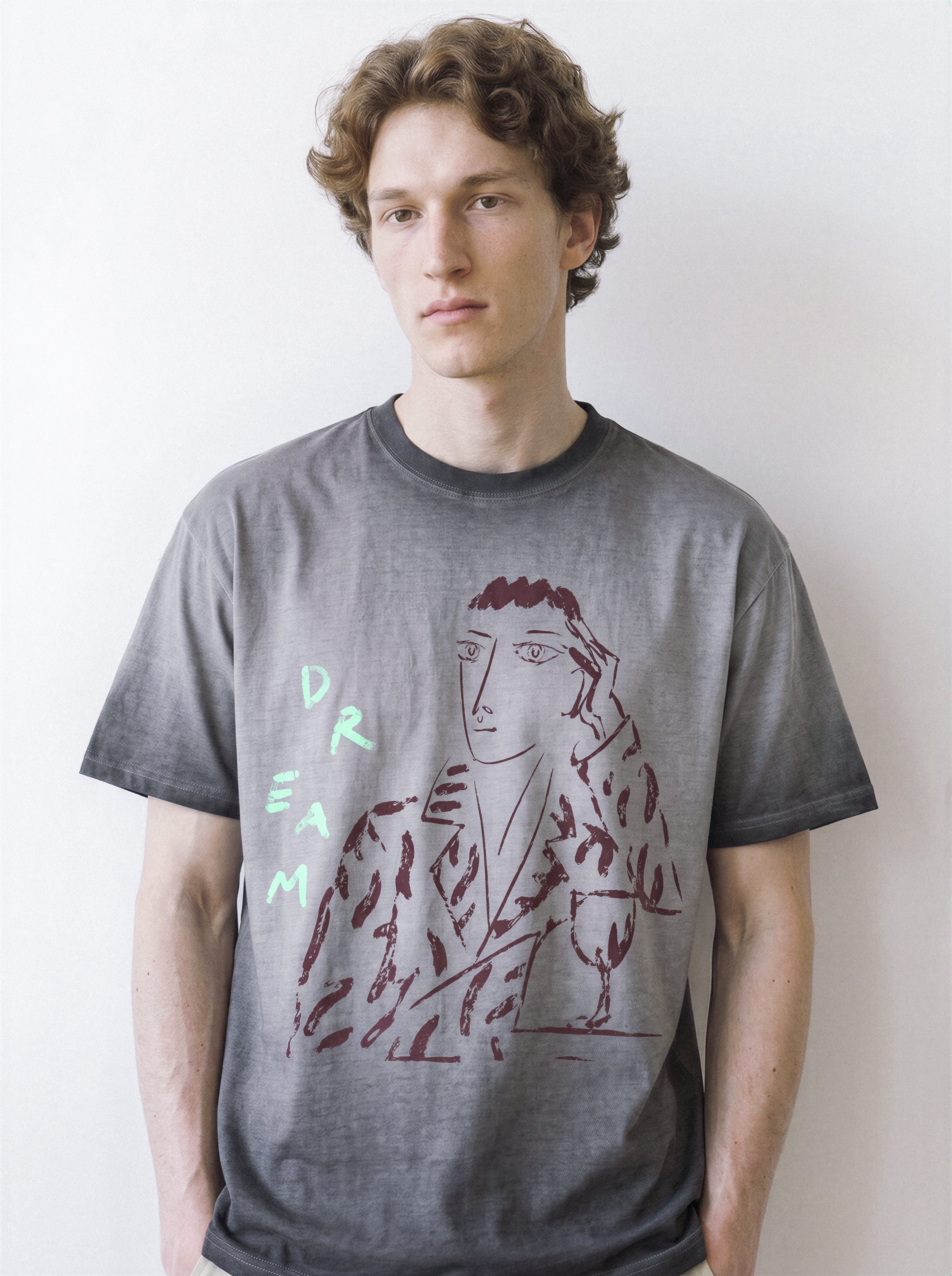 Front view of a male model standing naturally, demonstrating the structured drape of an ombre grey 230g premium cotton tee with a daydreamer charcoal sketch design.