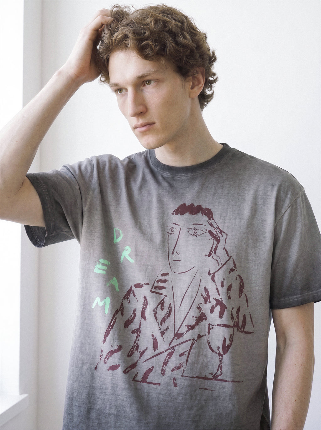 Front portrait of a male model with his hand in his hair, showcasing the fit of an ombre grey heavyweight boxy fit tee with a striking charcoal daydreamer illustration.