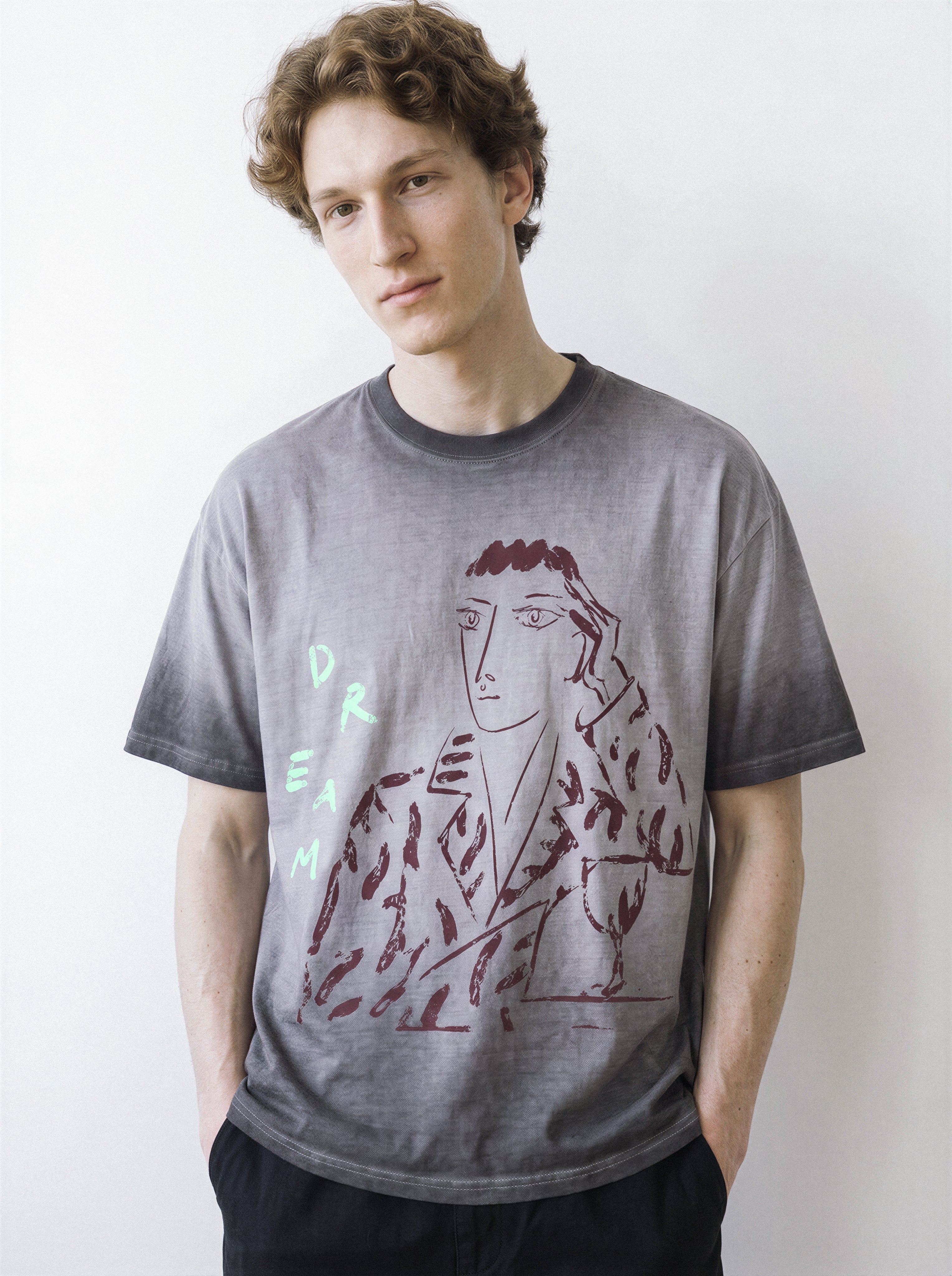 A male model in a relaxed stance, wearing an ombre grey structured streetwear t-shirt highlighting a modern charcoal portrait and bold green typography.