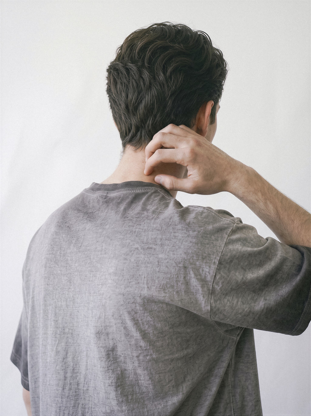 A man seen from behind wearing a heather gray short-sleeve t-shirt, touching the back of his neck, against a plain off-white background.