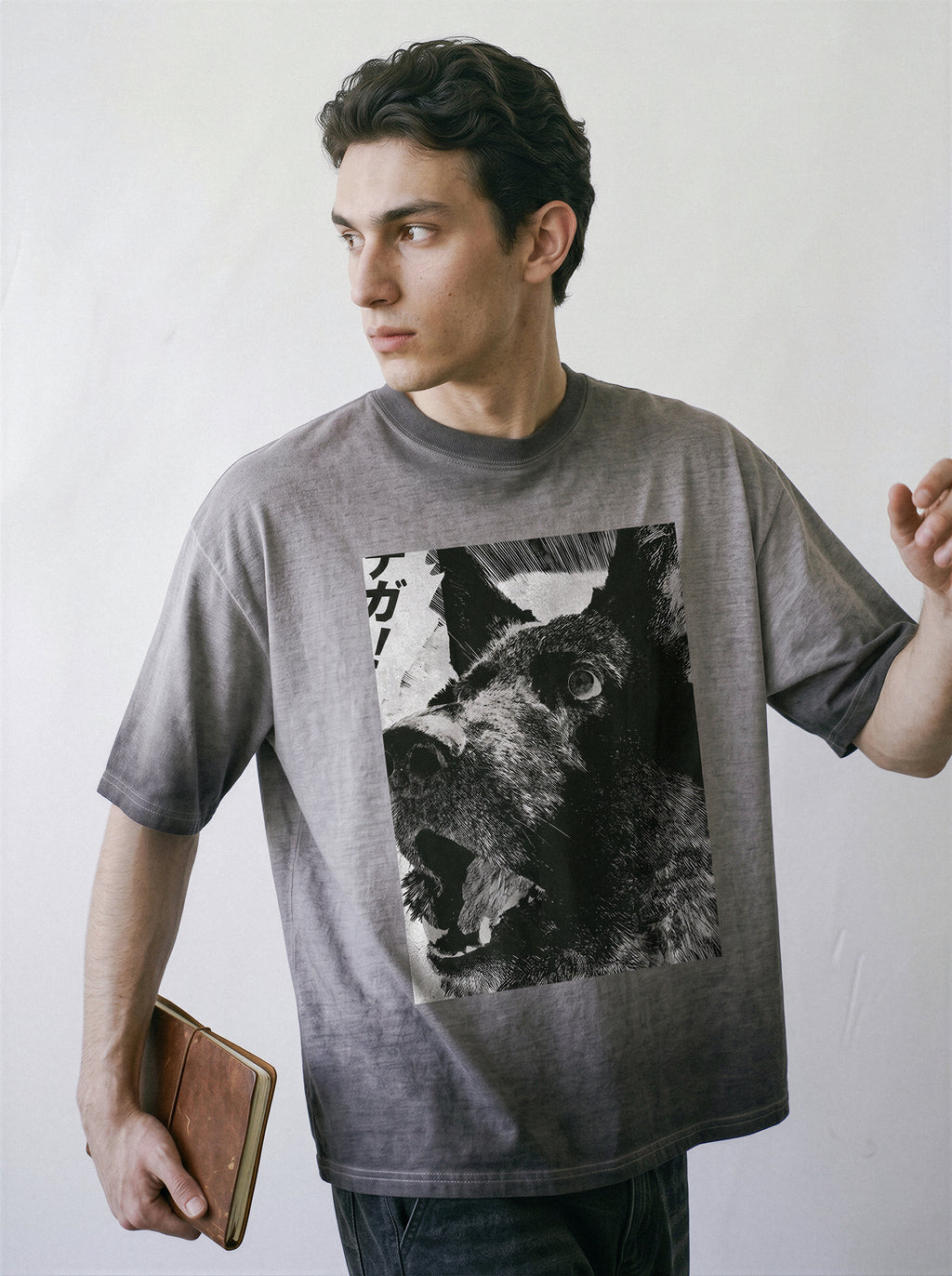 A male model holding a notebook and looking away, wearing an ombre grey structured streetwear t-shirt highlighting a monochrome line art dog illustration.