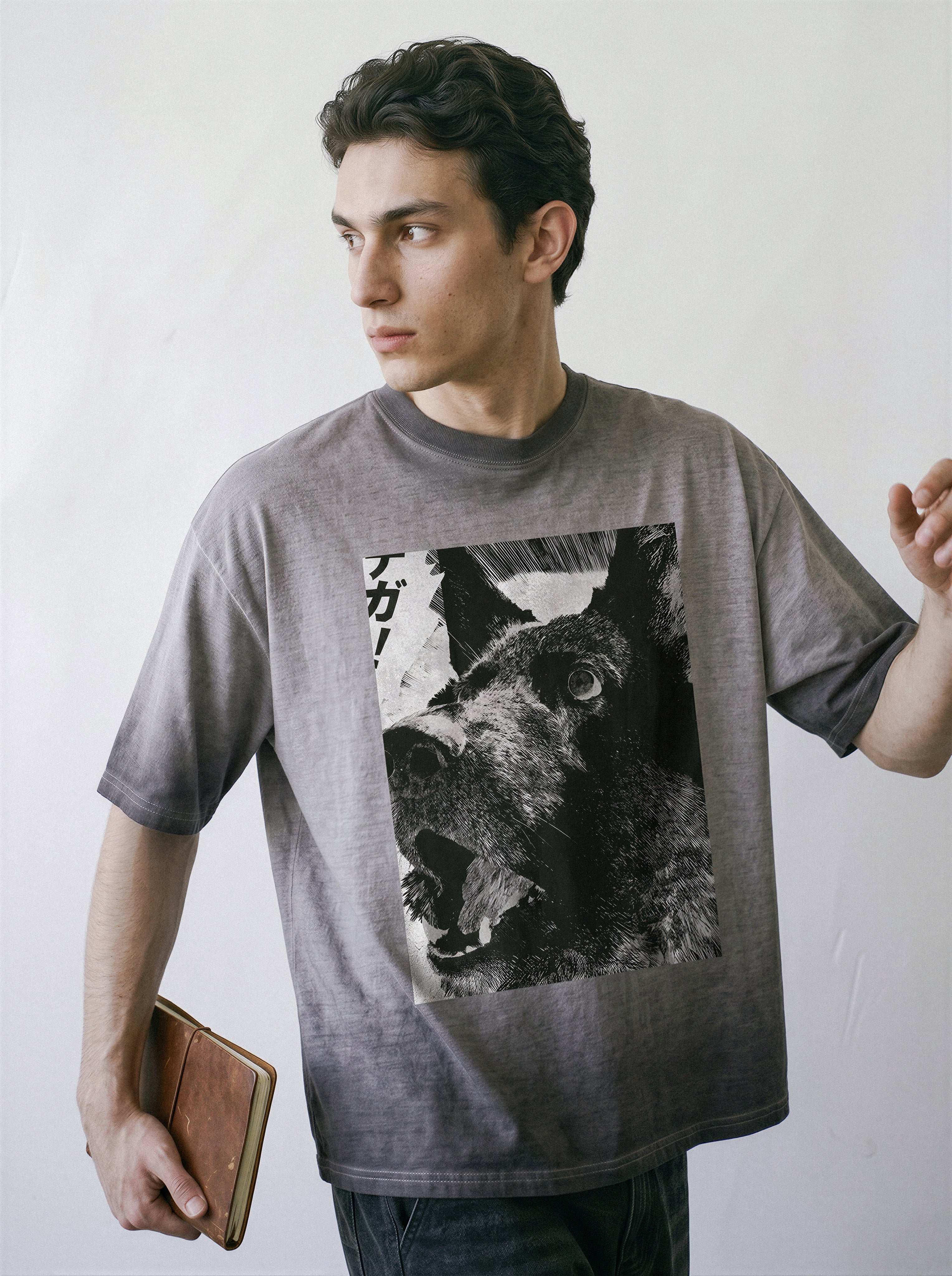 A male model holding a notebook and looking away, wearing an ombre grey structured streetwear t-shirt highlighting a monochrome line art dog illustration.