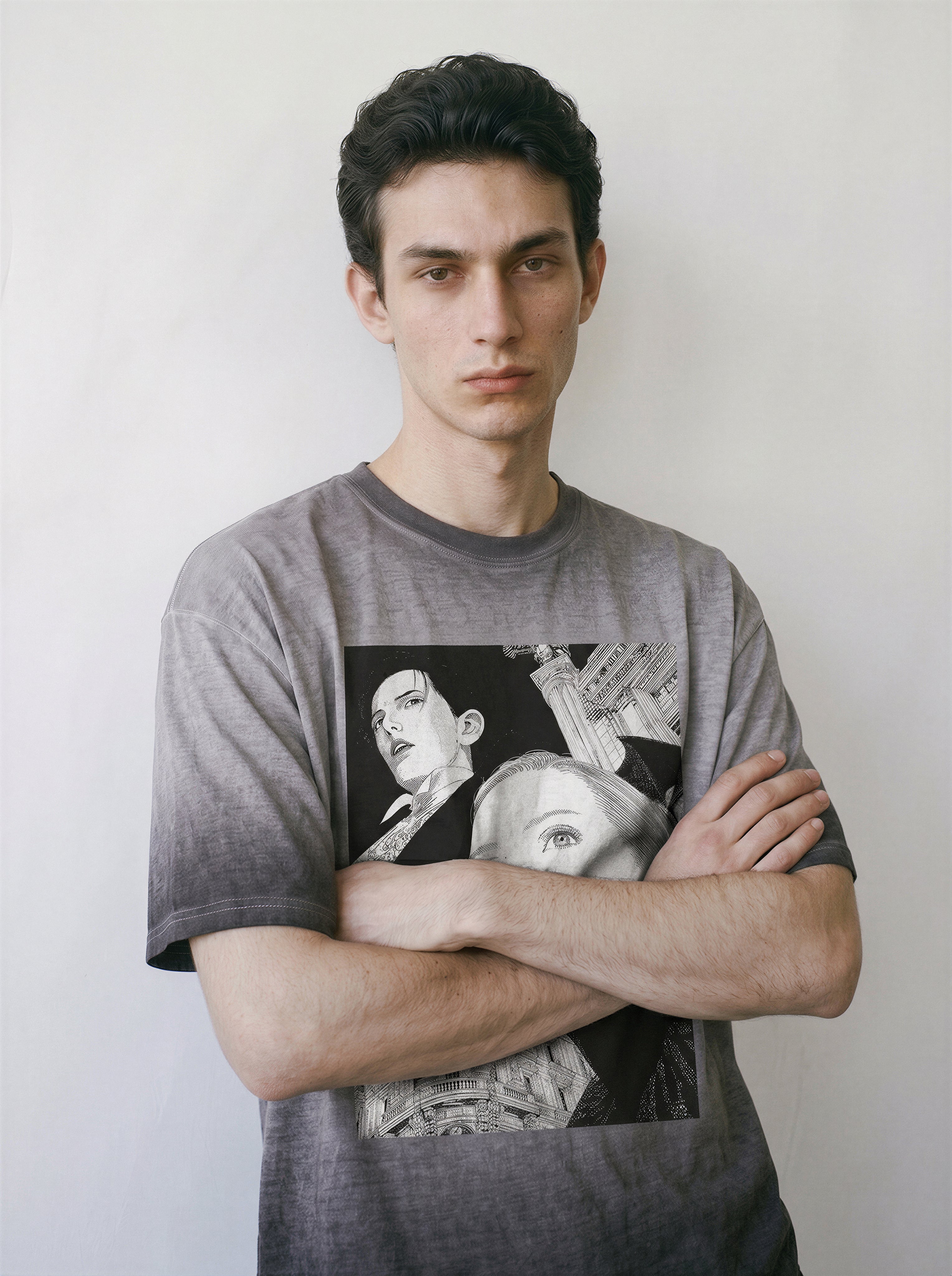 A young man with dark hair stands with arms crossed, wearing a gray short-sleeve t-shirt with a vintage manga generational shift design of two faces on the front, set against a plain off-white wall.