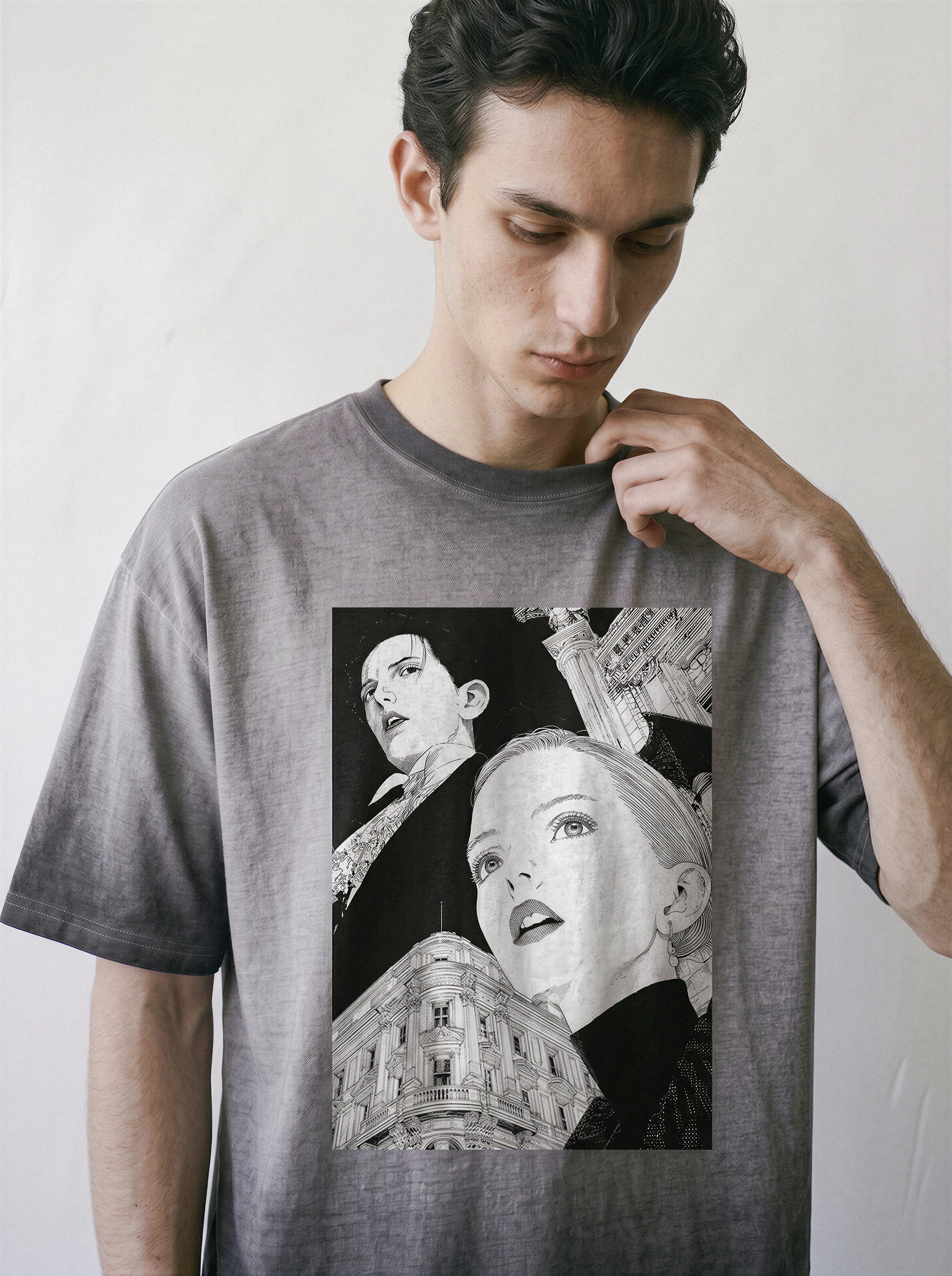 A man wearing a gray short-sleeve graphic t-shirt,featuring a monochrome retro manga architecture print of one man and a woman and a building, posed against a light gray wall.