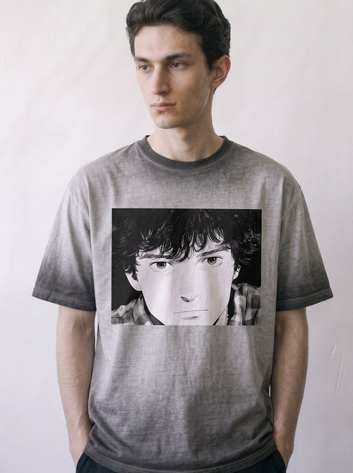 Close-up shot of a model looking down, showcasing the detailed retro manga portrait print on an ombre grey heavyweight boxy fit graphic tee.Showing a young man's face on the front, standing against a light gray background.