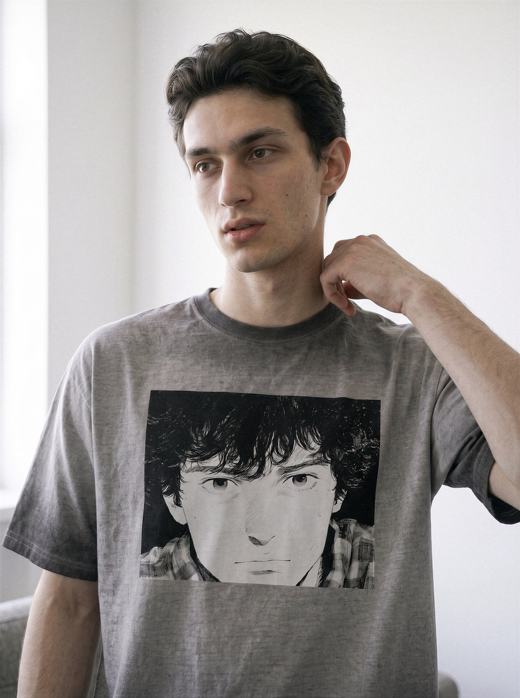 A man resting his hand on his neck, demonstrating the structured drape of an ombre grey heavyweight boxy fit t-shirt with a vintage manga graphic.Showing a face on the front, posed against a bright white indoor background.