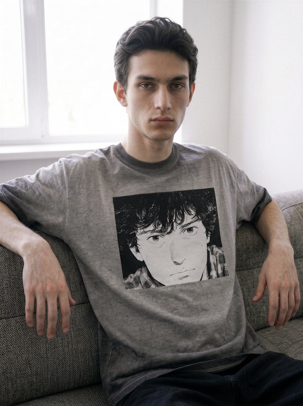 A male model sitting on a sofa, wearing an ombre grey structured streetwear t-shirt highlighting a 90s anime aesthetic monochrome comic print of a boy's face.