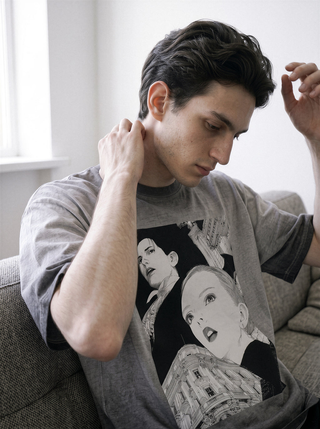 A man wearing a gray graphic t-shirt with a black and white illustration of a woman, sitting on a gray couch in a brightly lit room.