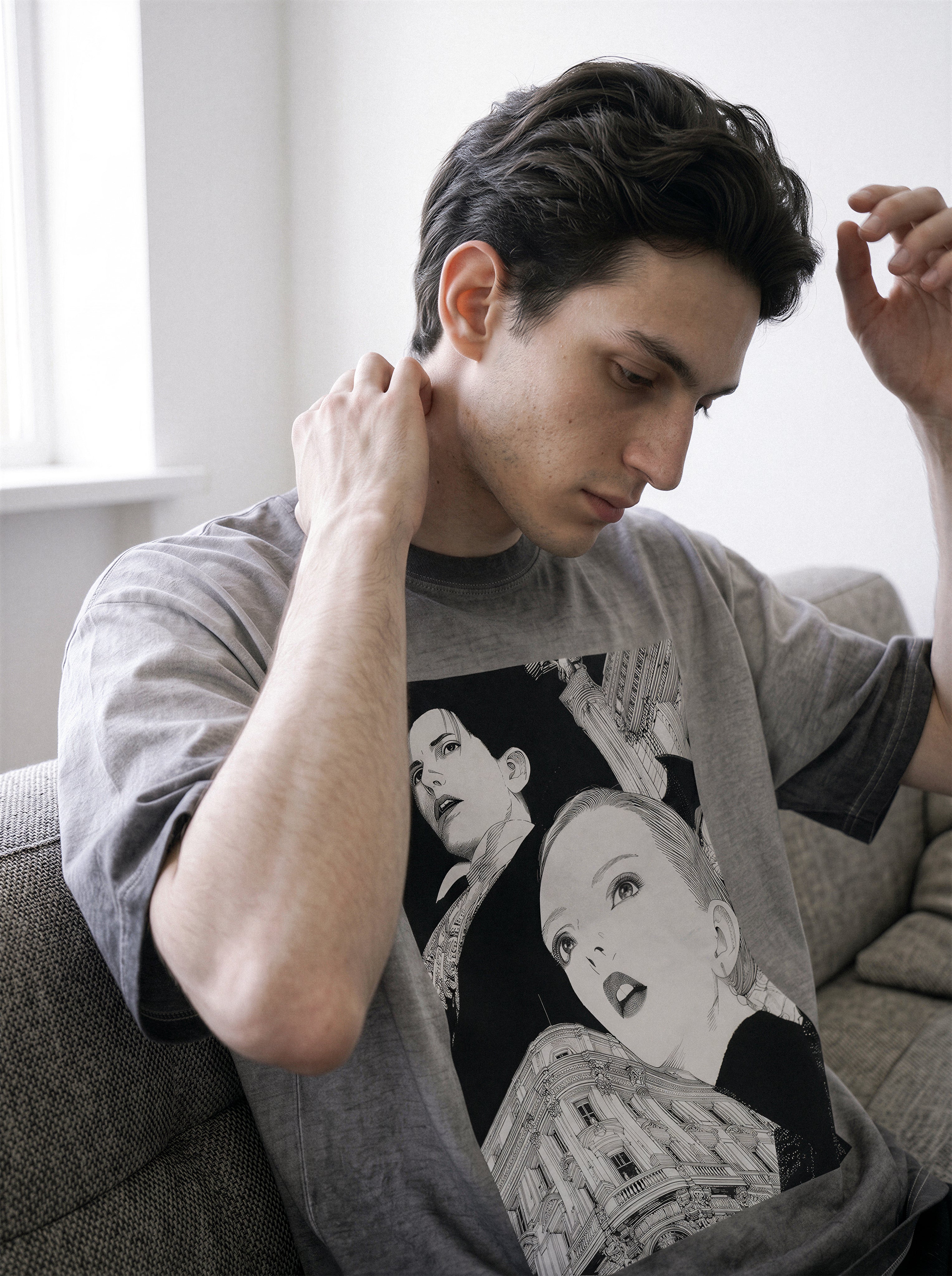 A man wearing a gray graphic t-shirt with a black and white illustration of a woman, sitting on a gray couch in a brightly lit room.