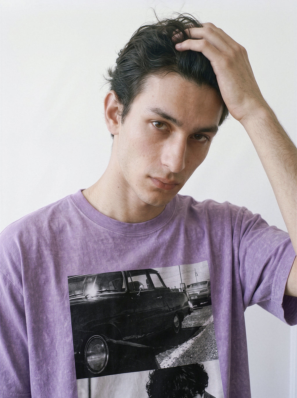 A male model with his hand in his hair looking down, showcasing the drape of an ombre violet structured streetwear tee with a hardboiled car and man split-panel graphic.