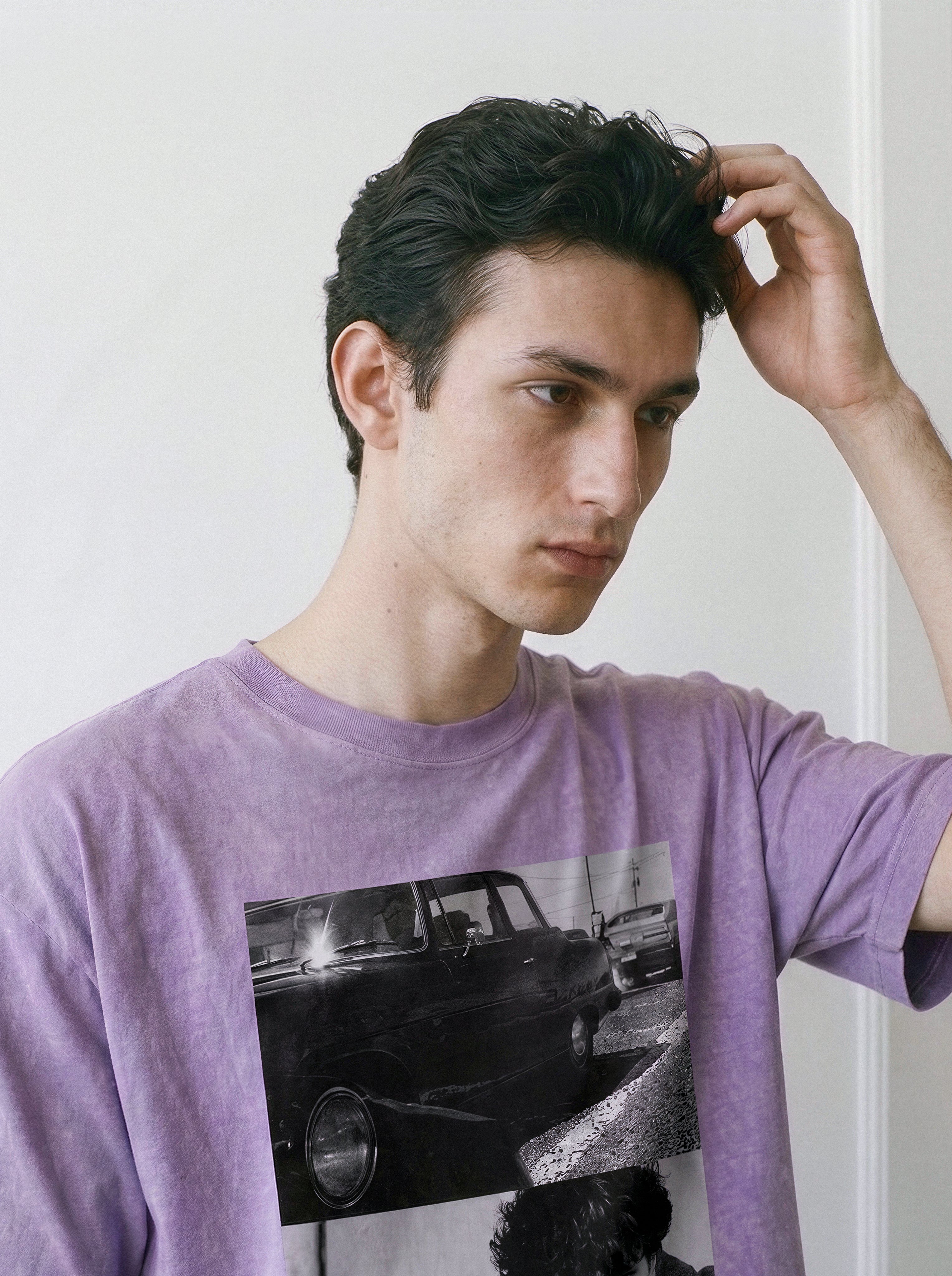 Model resting a hand on his head while looking away, wearing a premium ombre violet boxy fit t-shirt highlighting a 90s cinematic dual-panel photography print.