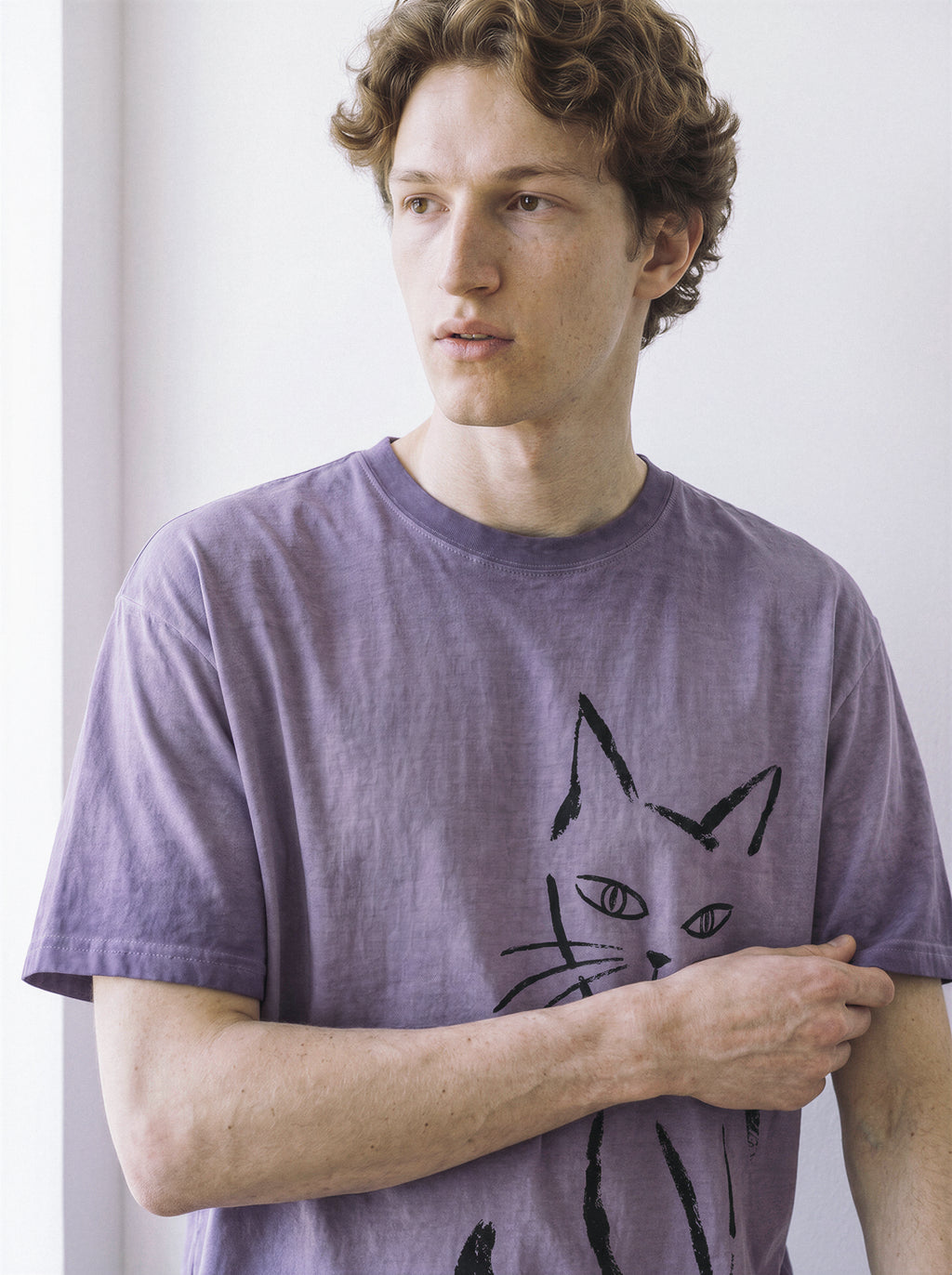 A male model looking to the side while adjusting the hem of an ombre violet structured streetwear t-shirt with a bold side-eye cat line art print.