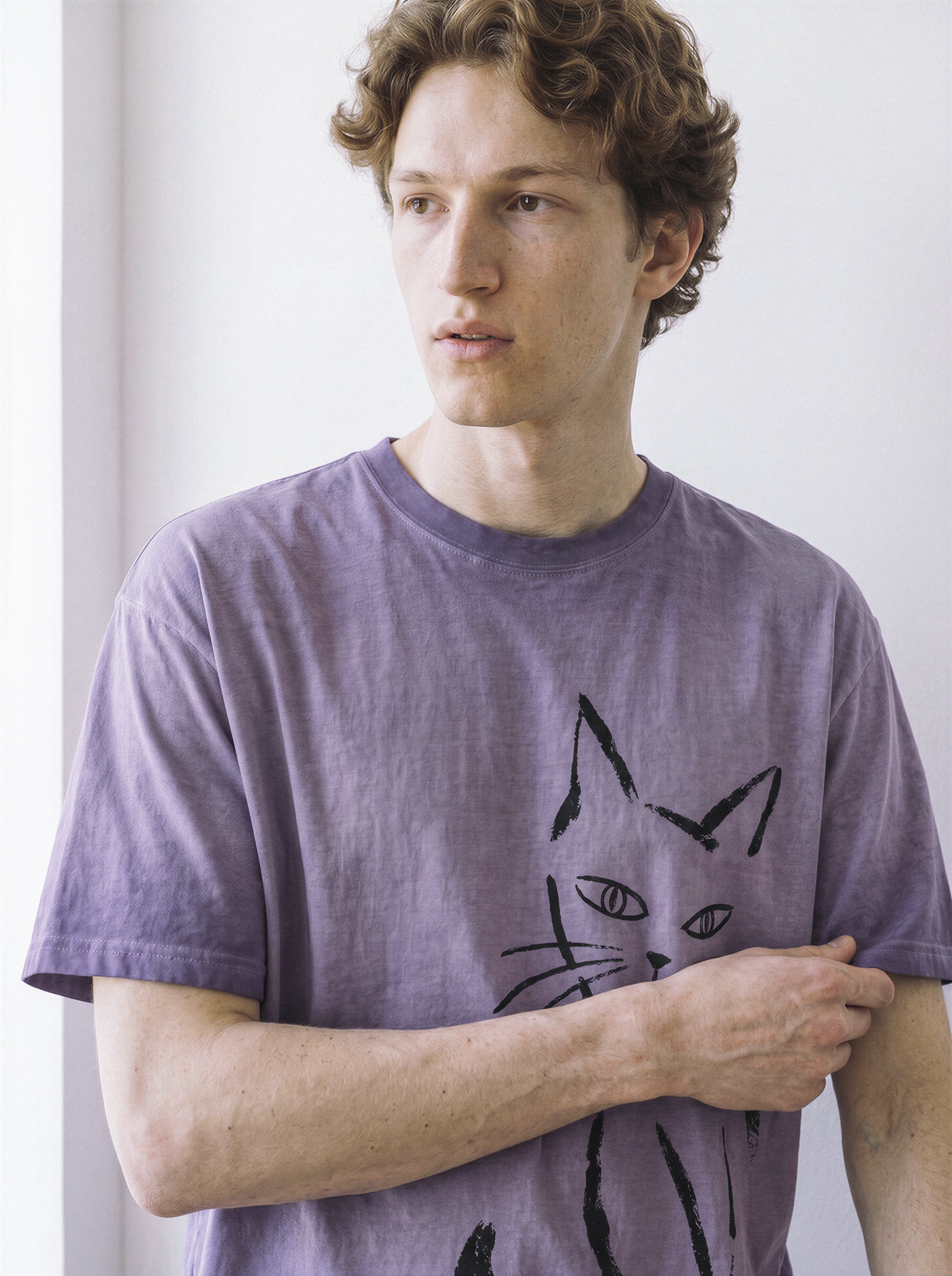 A male model looking to the side while adjusting the hem of an ombre violet structured streetwear t-shirt with a bold side-eye cat line art print.
