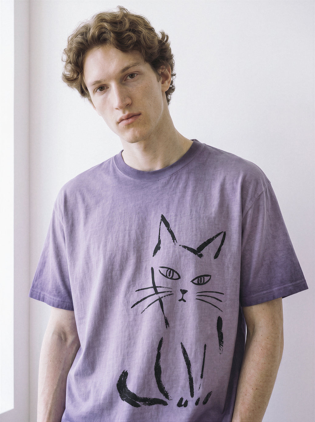 Front portrait of a male model looking down, wearing an ombre violet heavyweight boxy fit graphic tee featuring a raw paint stroke judgmental cat illustration.