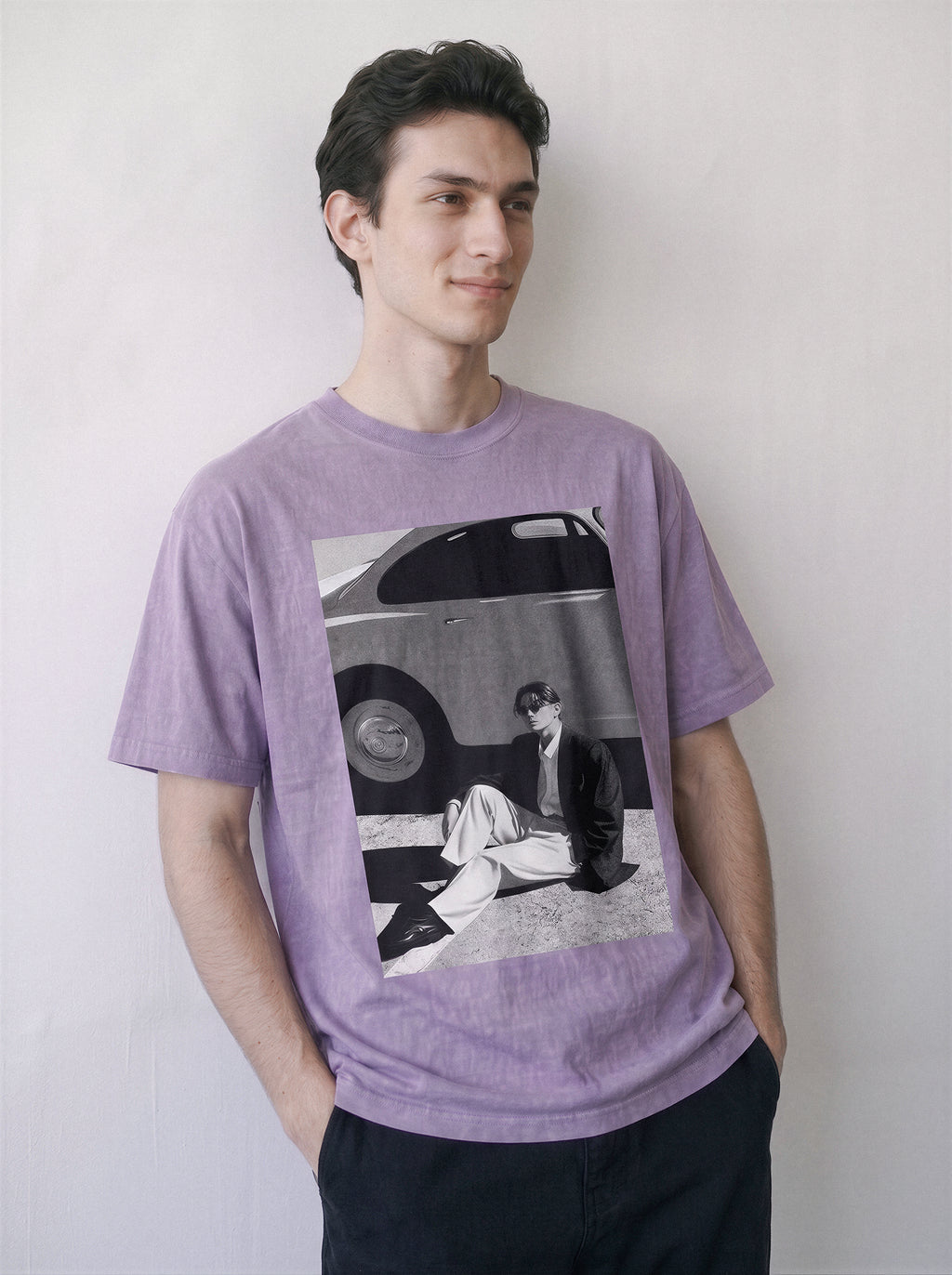 A man wearing a light purple heathered t-shirt with a large black and white graphic print on the front, posed against a plain off-white wall.