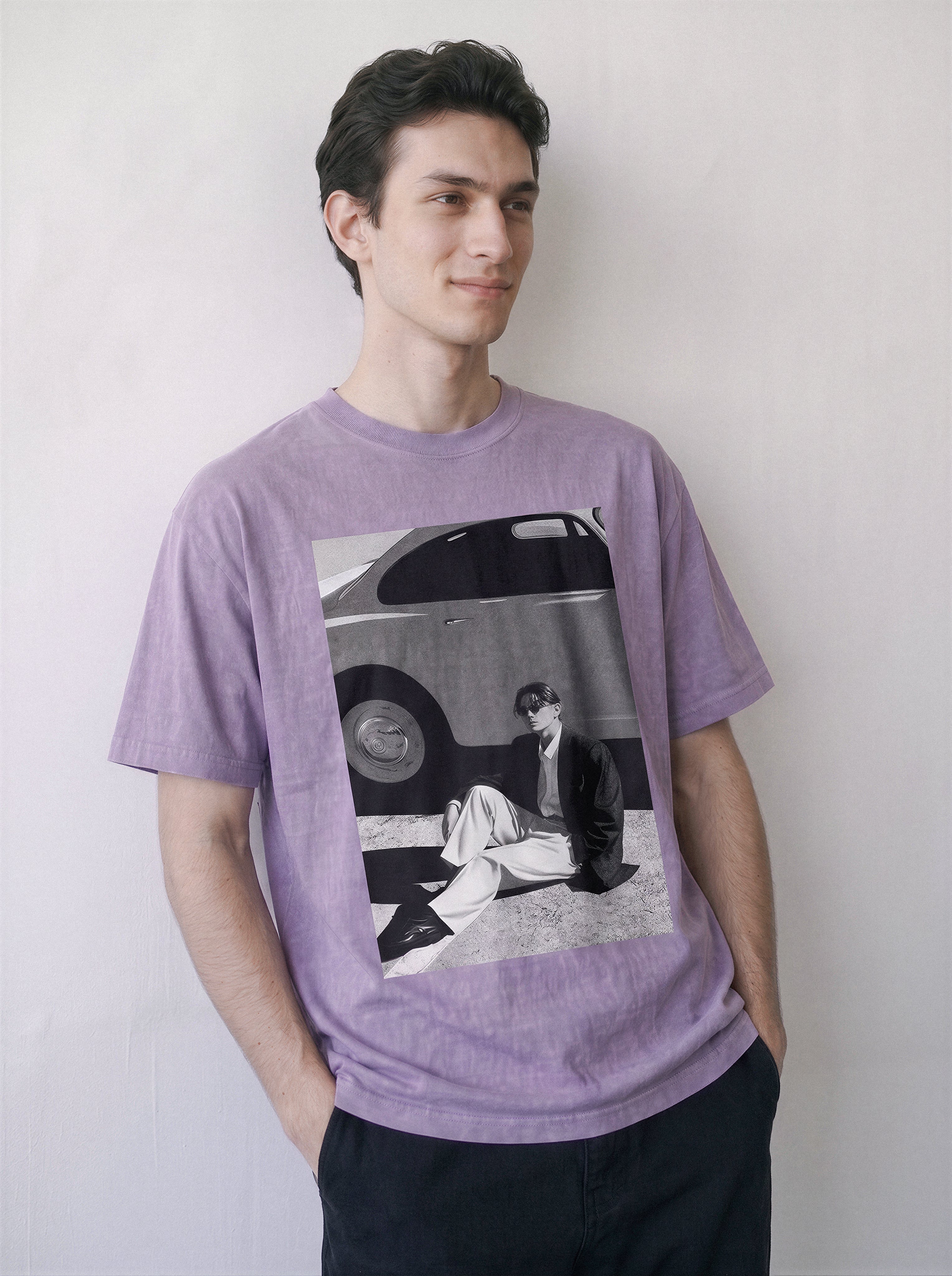 A man wearing a light purple heathered t-shirt with a large black and white graphic print on the front, posed against a plain off-white wall.