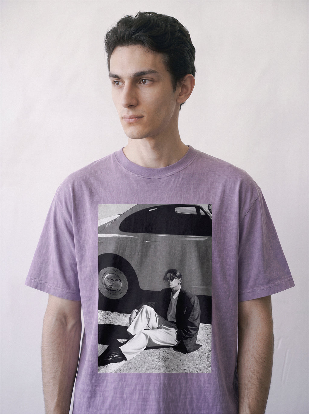 Close-up half-body shot of a male model looking away, wearing an ombre violet heavyweight boxy fit T-shirt with a 90s retro cinematic black and white photography print on the front showing a vintage car and a seated figure, posed against a light off-white background.