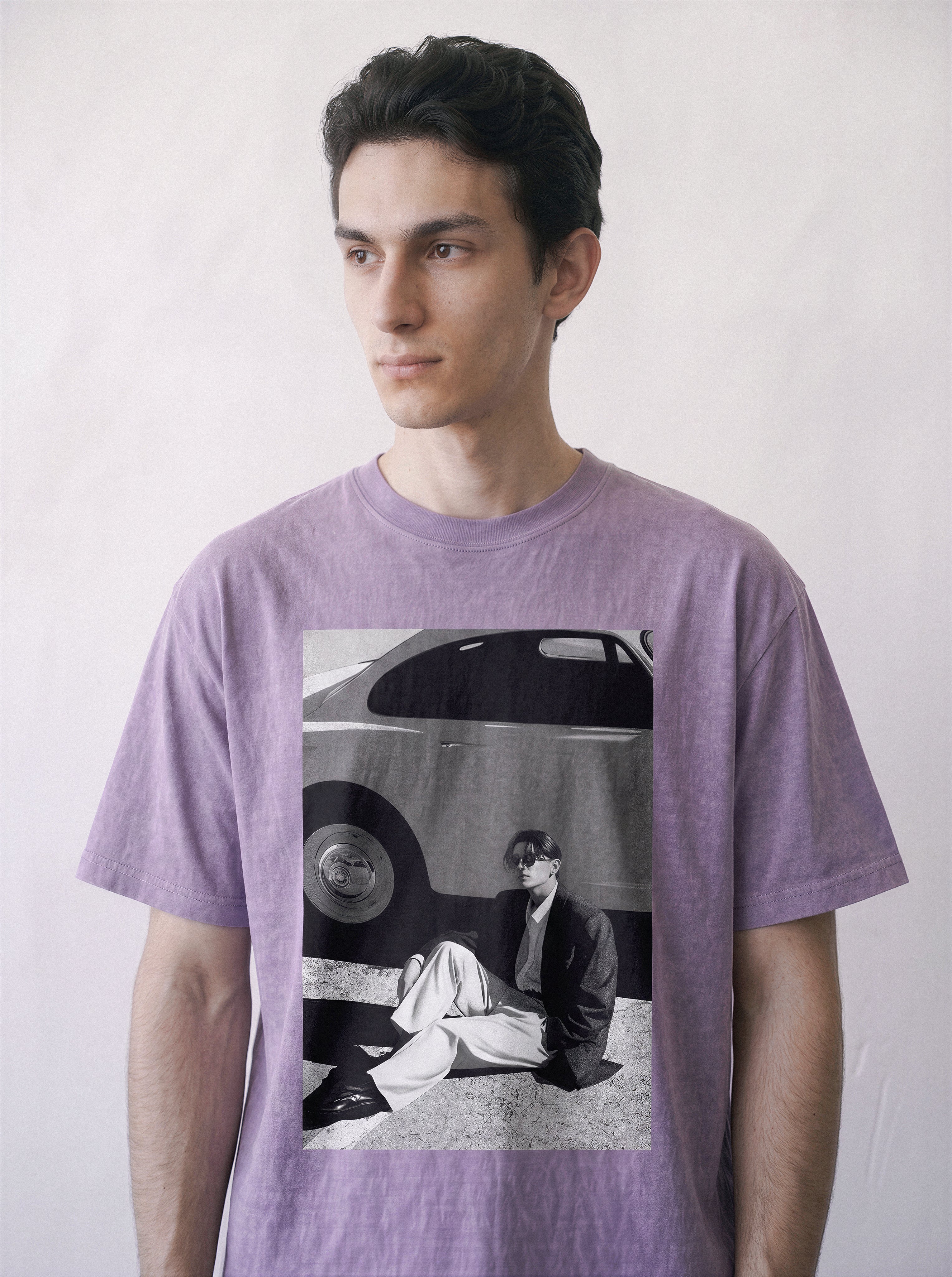 Close-up half-body shot of a male model looking away, wearing an ombre violet heavyweight boxy fit T-shirt with a 90s retro cinematic black and white photography print on the front showing a vintage car and a seated figure, posed against a light off-white background.