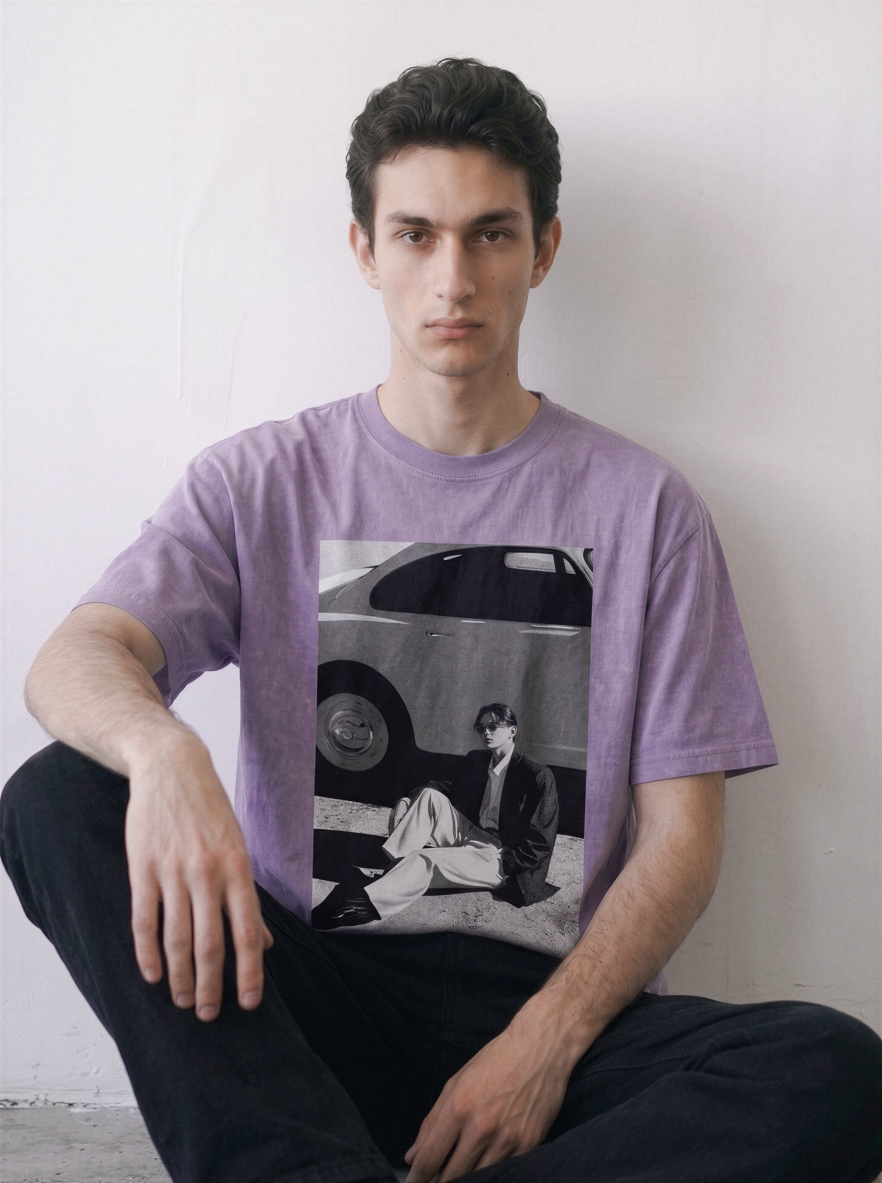 A man wearing an ombre violet heavyweight boxy fit graphic tee featuring a retro 90s photography print of a vintage car and a seated figure, paired with black pants, sitting against a plain off-white wall.