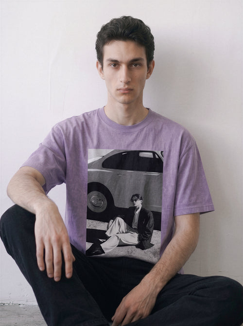A man wearing an ombre violet heavyweight boxy fit graphic tee featuring a retro 90s photography print of a vintage car and a seated figure, paired with black pants, sitting against a plain off-white wall.