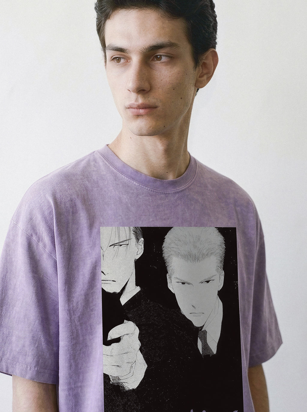 A man wearing a lavender-purple oversized t-shirt with a monochrome retro manga syndicate hitmen print on the front, set against a plain off-white background.