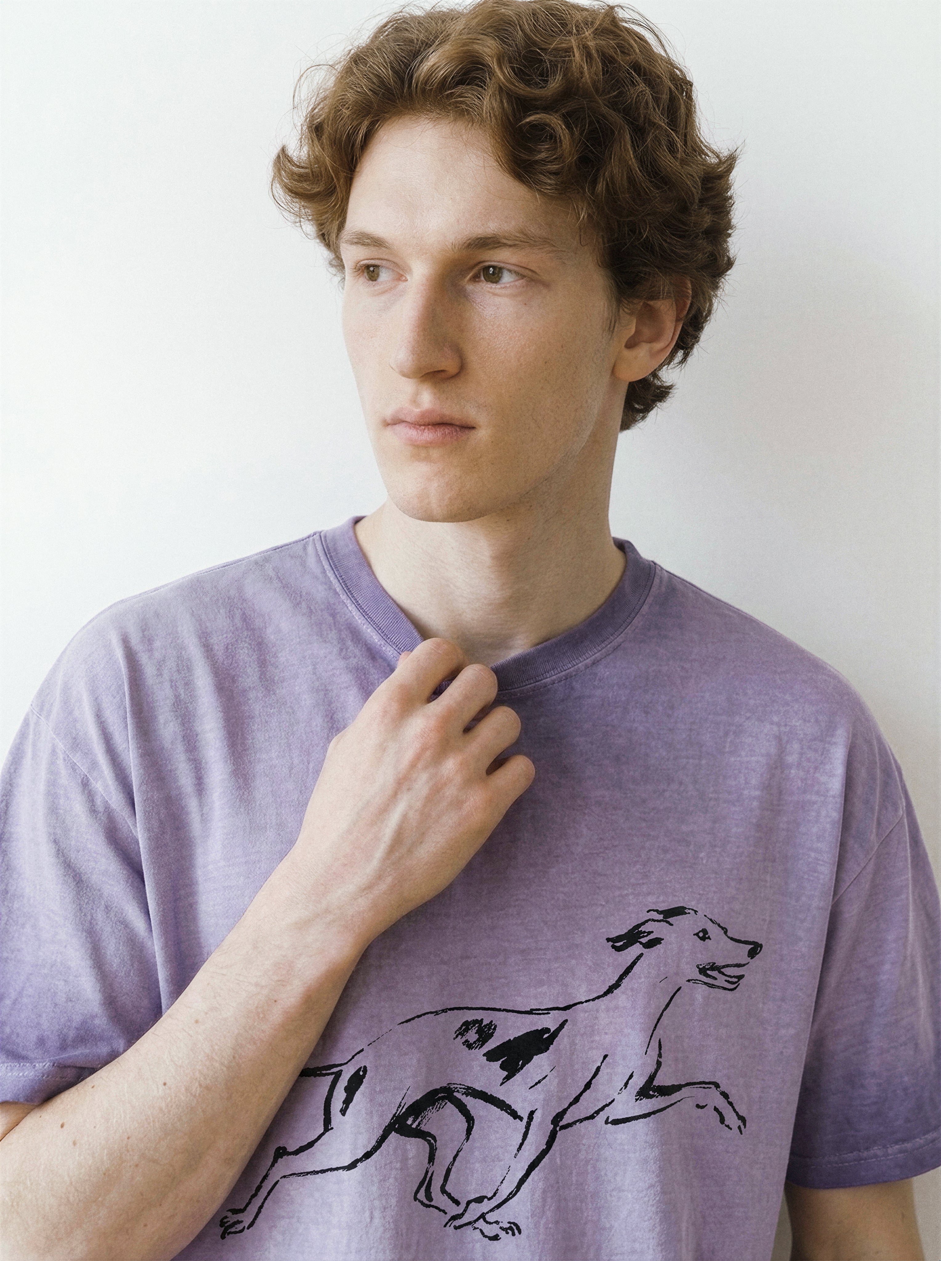 Front portrait of a male model adjusting the collar of an ombre violet graphic tee, featuring a minimalist running dog line art illustration.