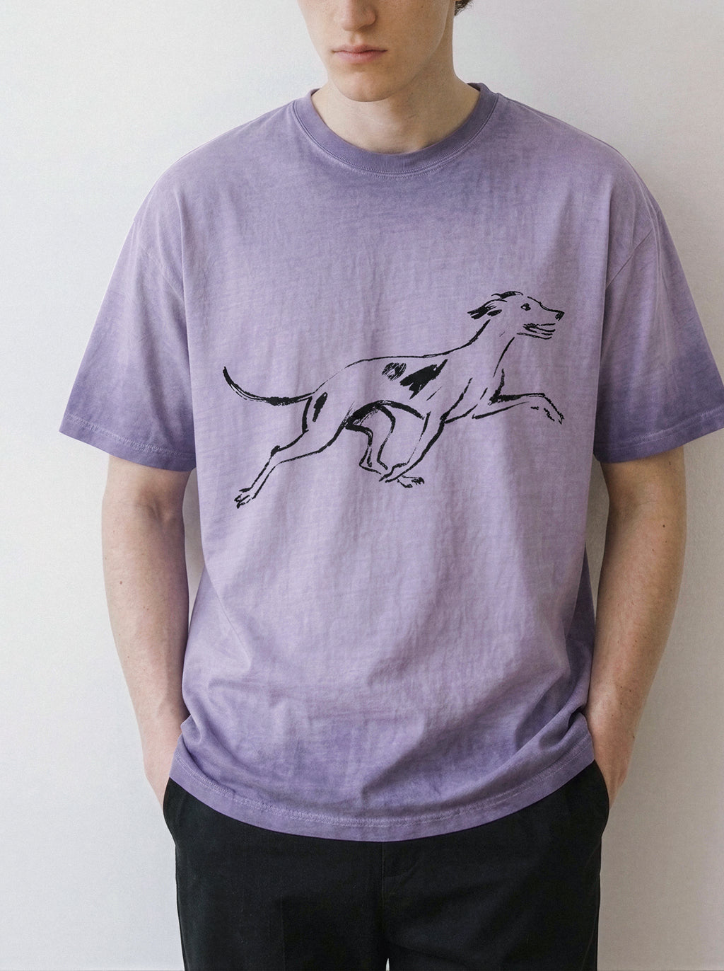 A person wearing a light purple crewneck t-shirt with a black line art graphic of a running dog on the front, paired with black pants, against a plain off-white wall.