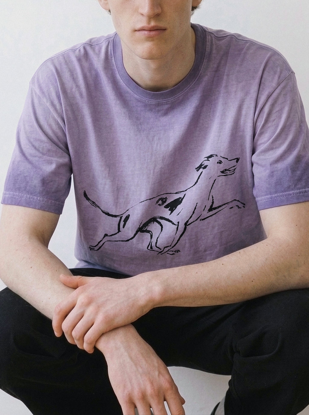 A male model leaning forward, showcasing the relaxed drape of an ombre violet t-shirt highlighting a pure line art running dog graphic.